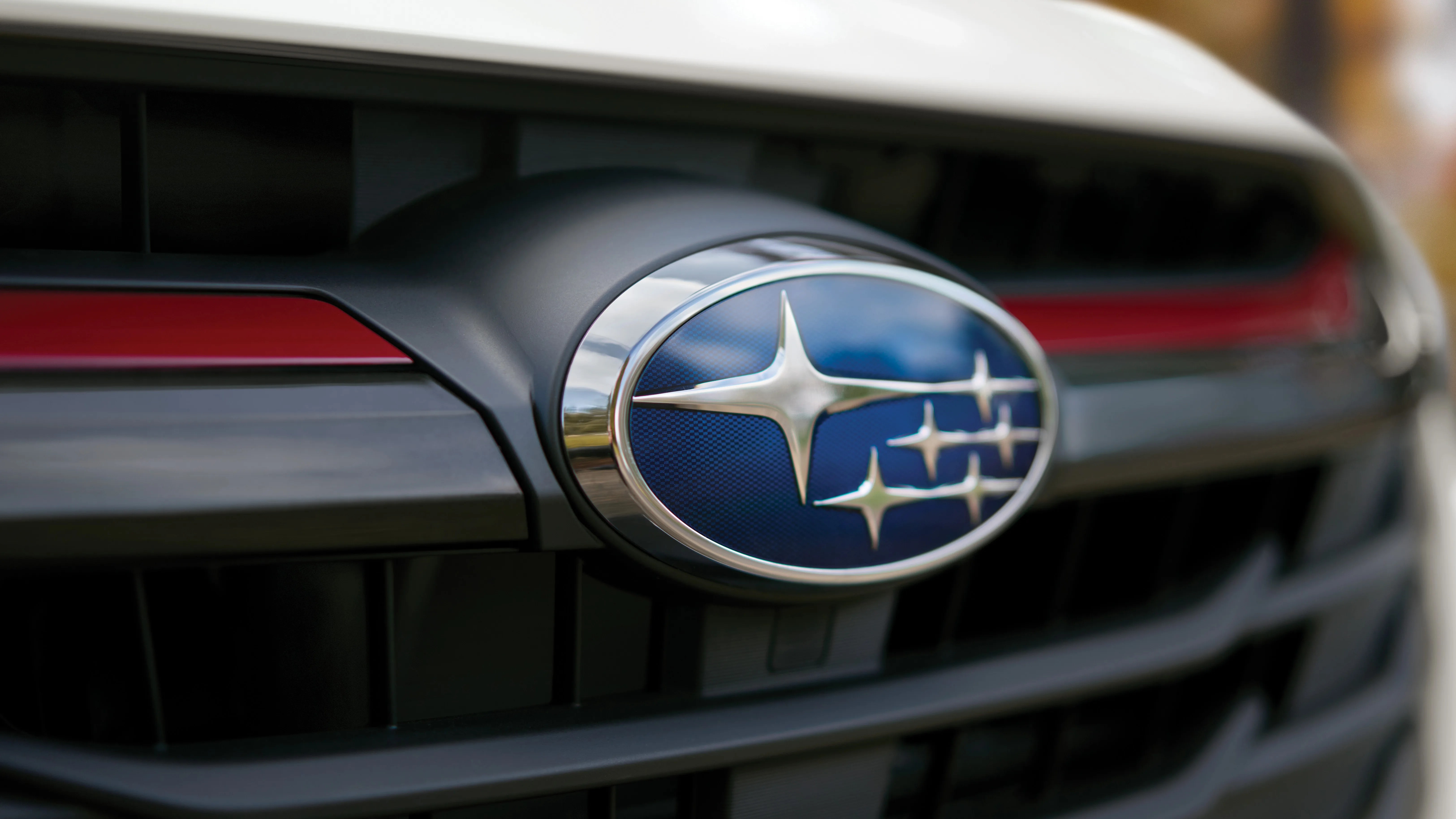 Close-up of a Subaru car emblem on the front grille, featuring a blue oval background with six silver stars and a chrome border, set against a black grille with a red accent line.
