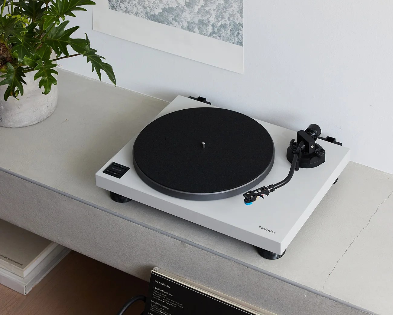 A white Technics turntable with a black platter mat and tonearm, placed on a light gray concrete surface. To the left, there is a potted plant with green leaves, and above the turntable, part of a framed artwork is visible on the white wall. Below the surface, there are some books or records partially visible.