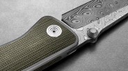 Close-up of a folding knife showing the textured green handle with a circular metal pivot screw and a patterned Damascus steel blade with intricate swirling designs. The knife is placed on a gray surface.