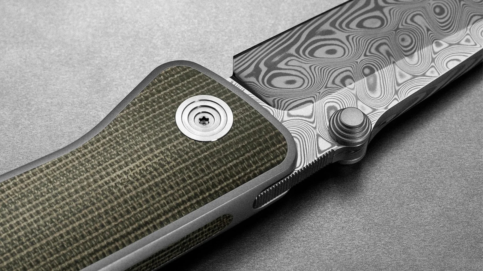 Close-up of a folding knife showing the textured green handle with a circular metal pivot screw and a patterned Damascus steel blade with intricate swirling designs. The knife is placed on a gray surface.