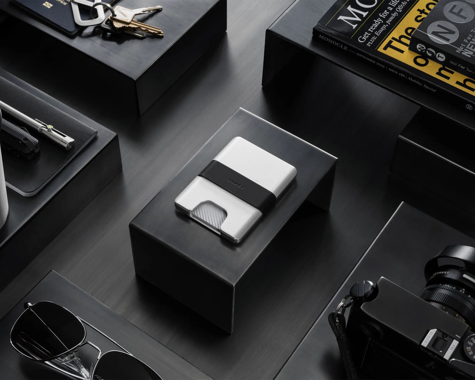 A sleek, minimalist silver and black wallet with the brand name "JAMES" embossed on the black band, placed on a black elevated platform. Surrounding it are various items including a pair of black sunglasses, a black camera with a lens, a set of keys with a keychain, a pen, a black notebook, and a magazine titled "MONOCLE." The overall setting is modern and monochromatic with a dark, matte surface.