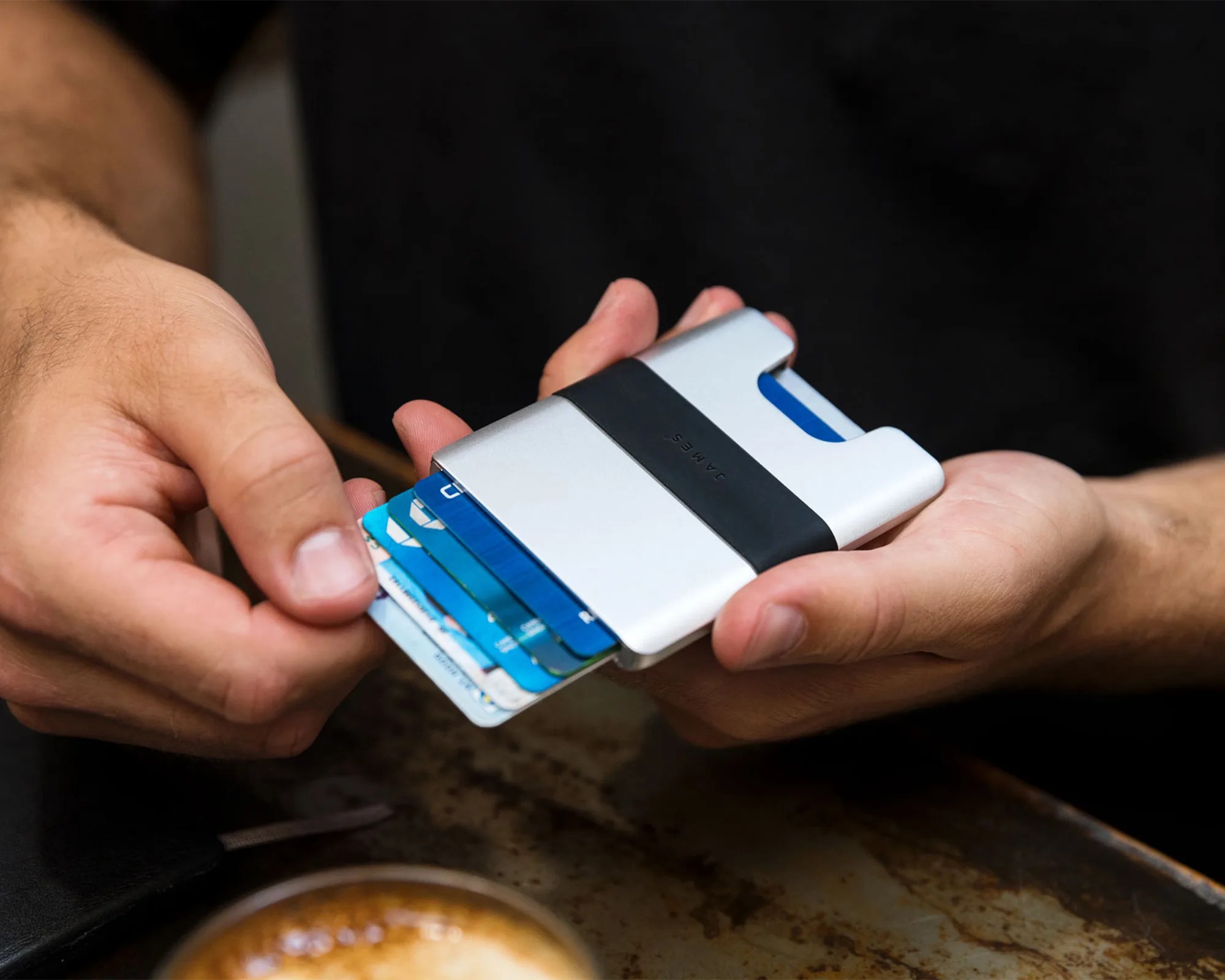 A person holding a sleek, silver and black minimalist wallet with several blue and white cards partially pulled out from it. The wallet has the brand name "JAMES" embossed on the black band. The background includes a blurred surface and a cup of coffee.