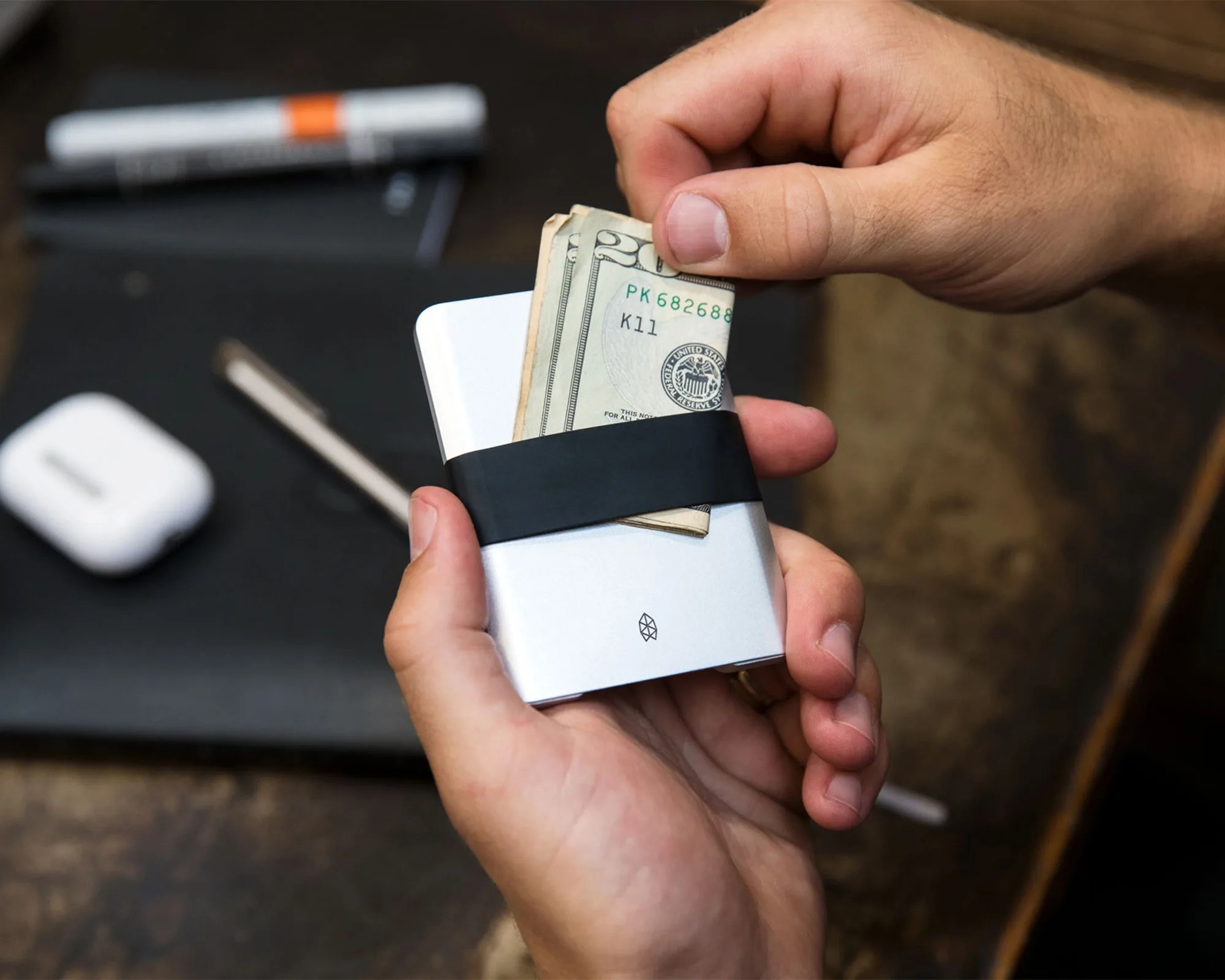 Hands holding a slim, silver metal wallet with a black elastic band securing folded US dollar bills. The wallet features a small geometric logo on the front. In the background, a blurred desk surface with a white wireless earbud case, a pen, and a marker are visible.