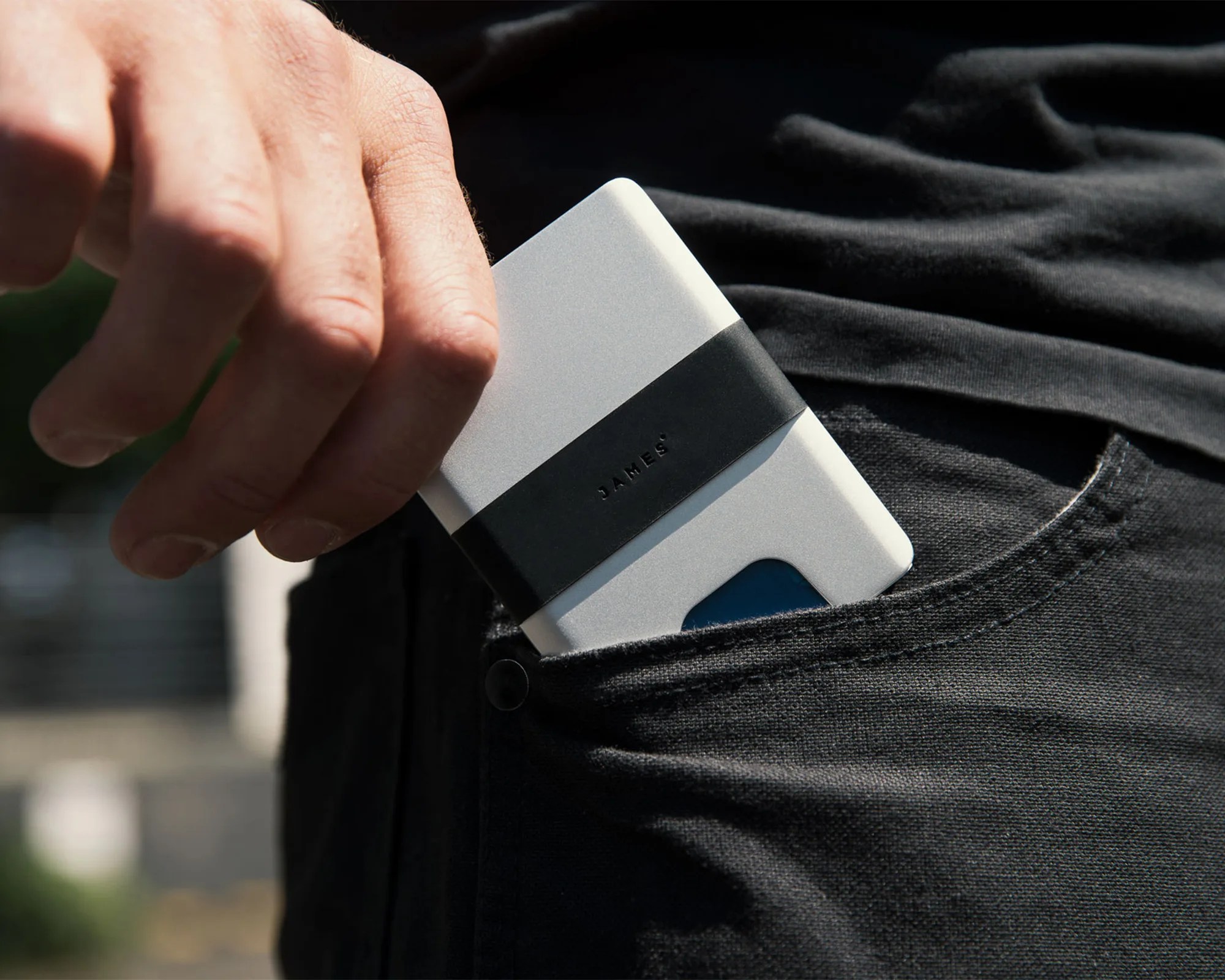 A hand is placing a slim, silver cardholder with a black band labeled "JAMES" into the front pocket of black pants. A blue card is partially visible in the cardholder.
