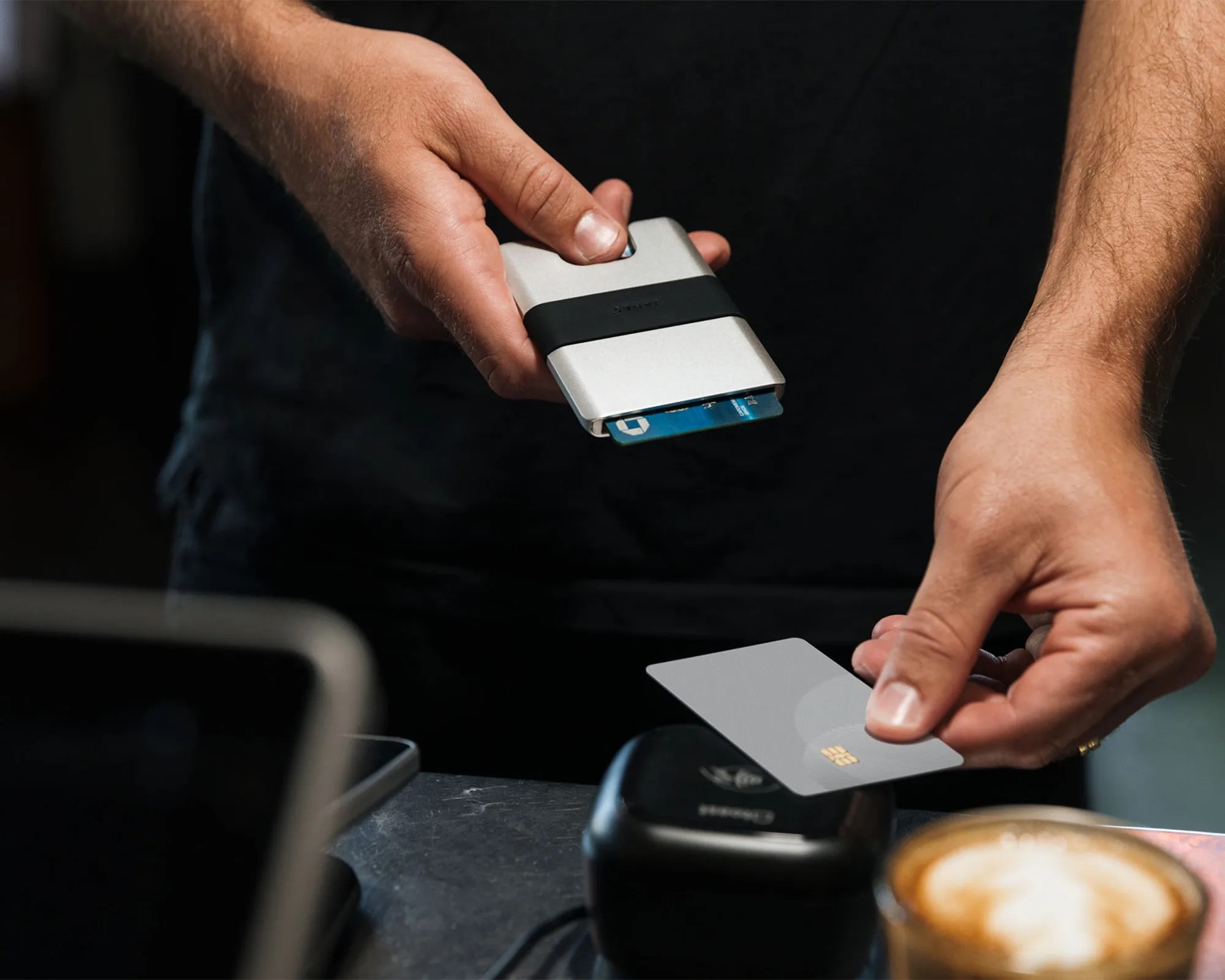 A person holding a slim, silver cardholder with a black band, partially revealing blue cards inside, while the other hand holds a gray chip-enabled card above a black card reader on a table. A cup of coffee with latte art is also visible in the foreground.