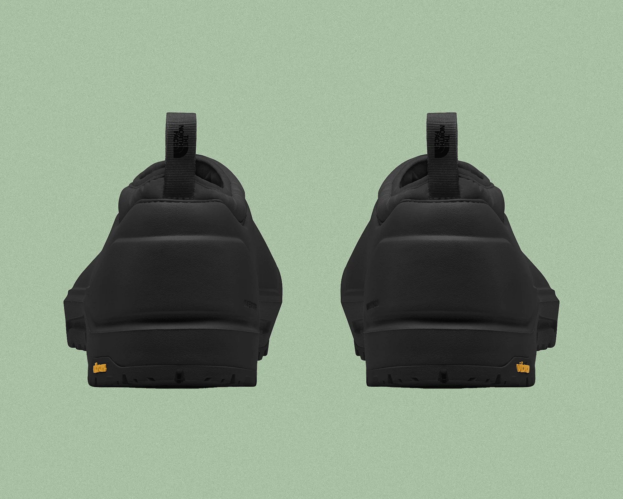 Pair of black slip-on shoes viewed from the back, featuring thick soles with a small yellow Vibram logo on each heel. Each shoe has a black pull tab with The North Face logo printed in black. The shoes have a smooth, matte finish and a minimalist design.