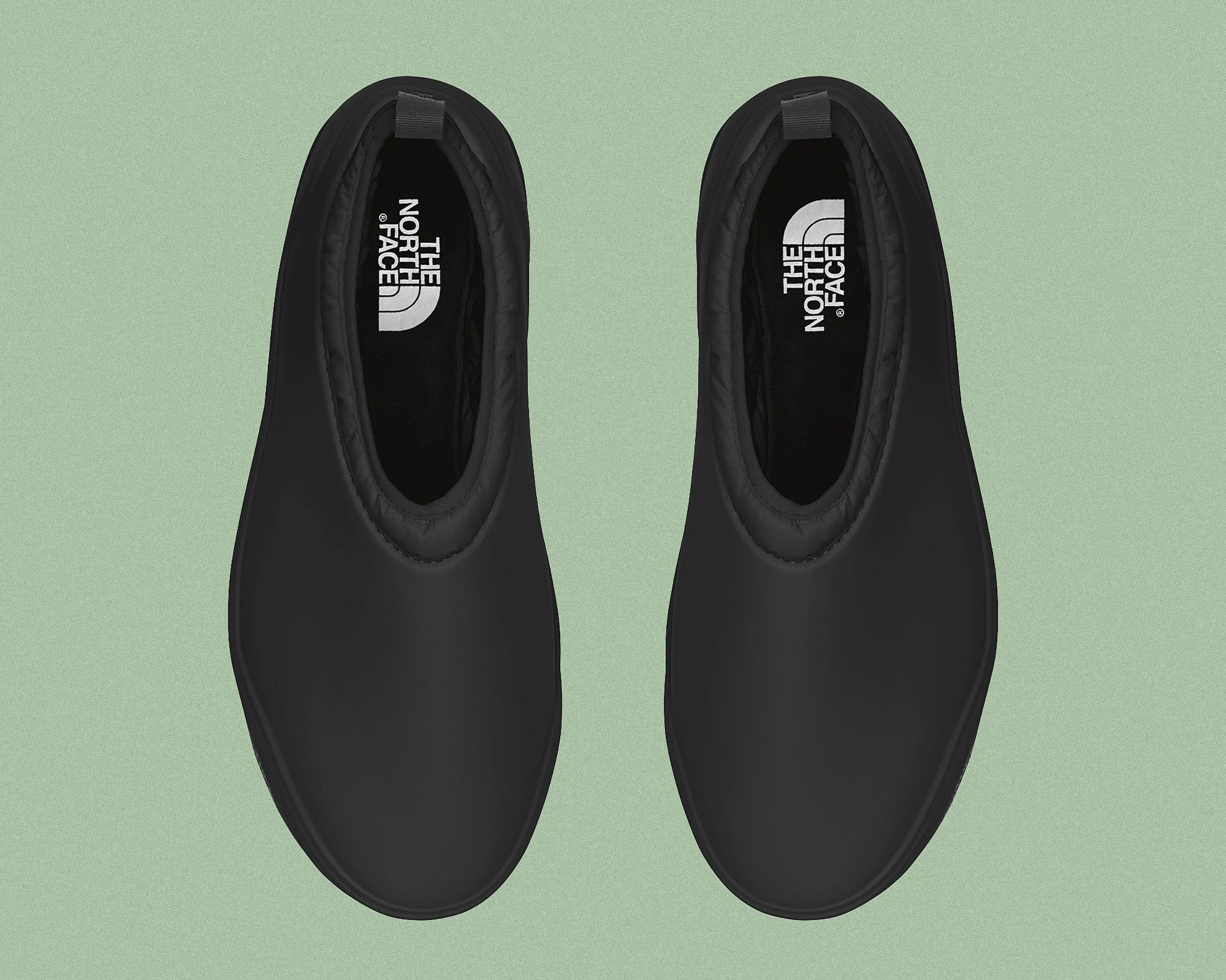 A pair of black slip-on shoes with a smooth, matte finish. The shoes have a rounded toe and a padded collar around the ankle. Inside the shoes, the insoles feature the white "The North Face" logo. The shoes are displayed from a top-down view on a light green background.