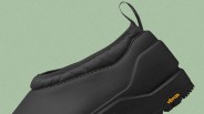 Black slip-on shoe with a padded collar and a pull tab at the heel, featuring a thick sole with a Vibram logo in yellow on the side. The shoe has a smooth, matte finish and a minimalist design.