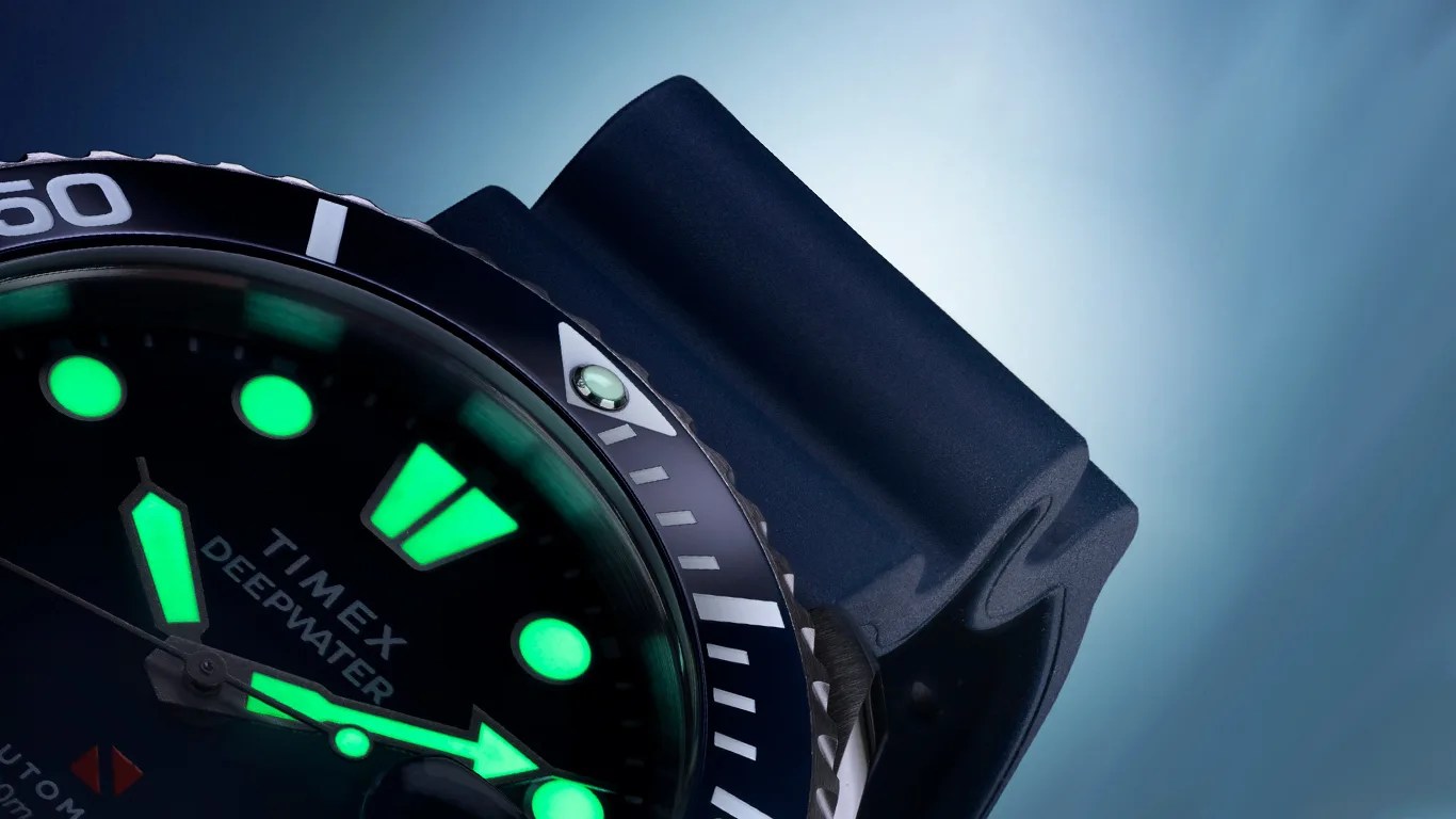 Close-up of a Timex Deepwater dive watch face with glowing green hour markers and hands, a black bezel with white markings, and a dark blue strap.