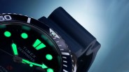 Close-up of a Timex Deepwater dive watch face with glowing green hour markers and hands, a black bezel with white markings, and a dark blue strap.