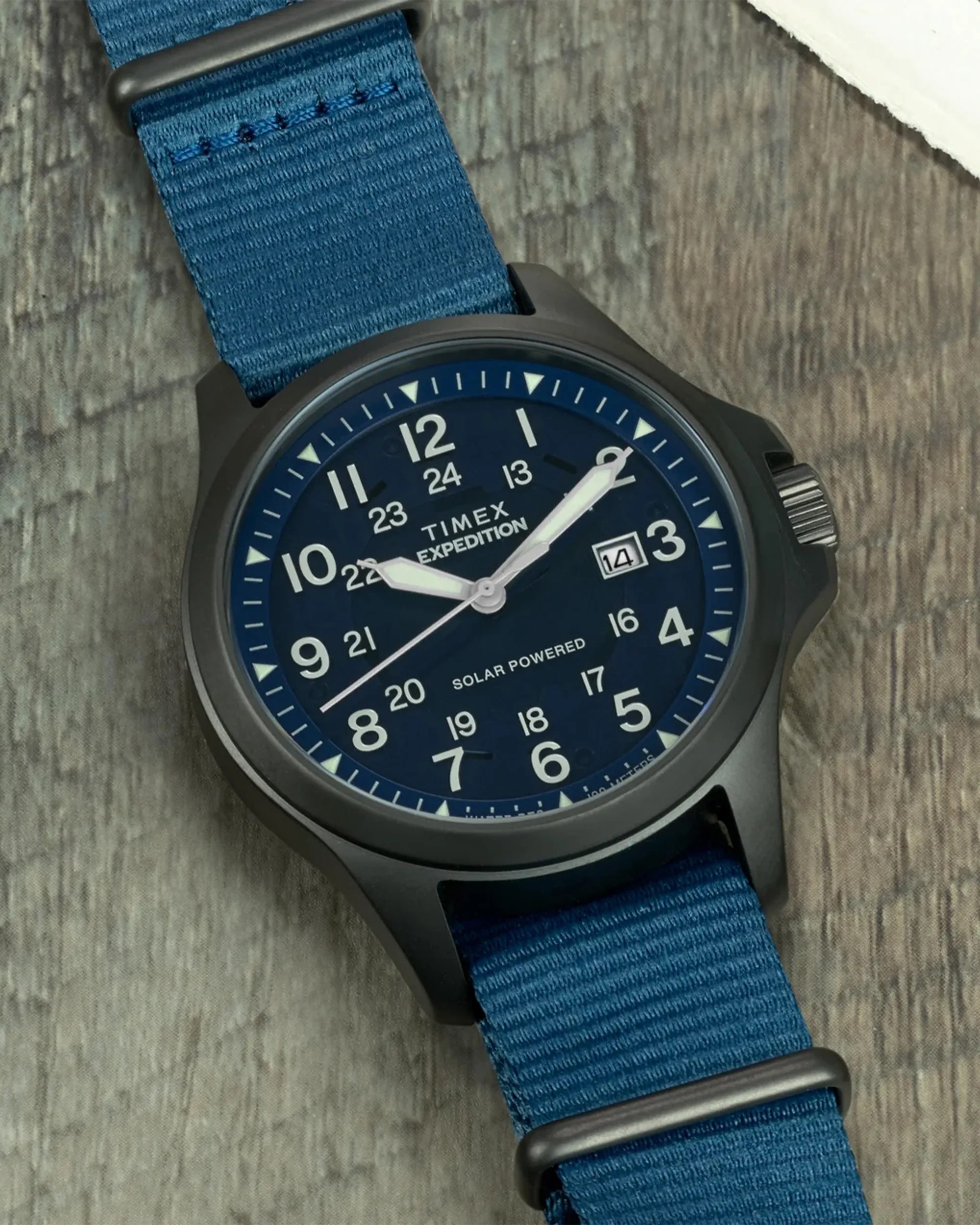 Timex Expedition wristwatch with a black case, dark blue dial, and white numerals. The watch features a date window at the 3 o'clock position, white hour and minute hands, and a blue fabric strap with black hardware. The dial also includes 24-hour military time markings and the text "TIMEX EXPEDITION" and "SOLAR POWERED.