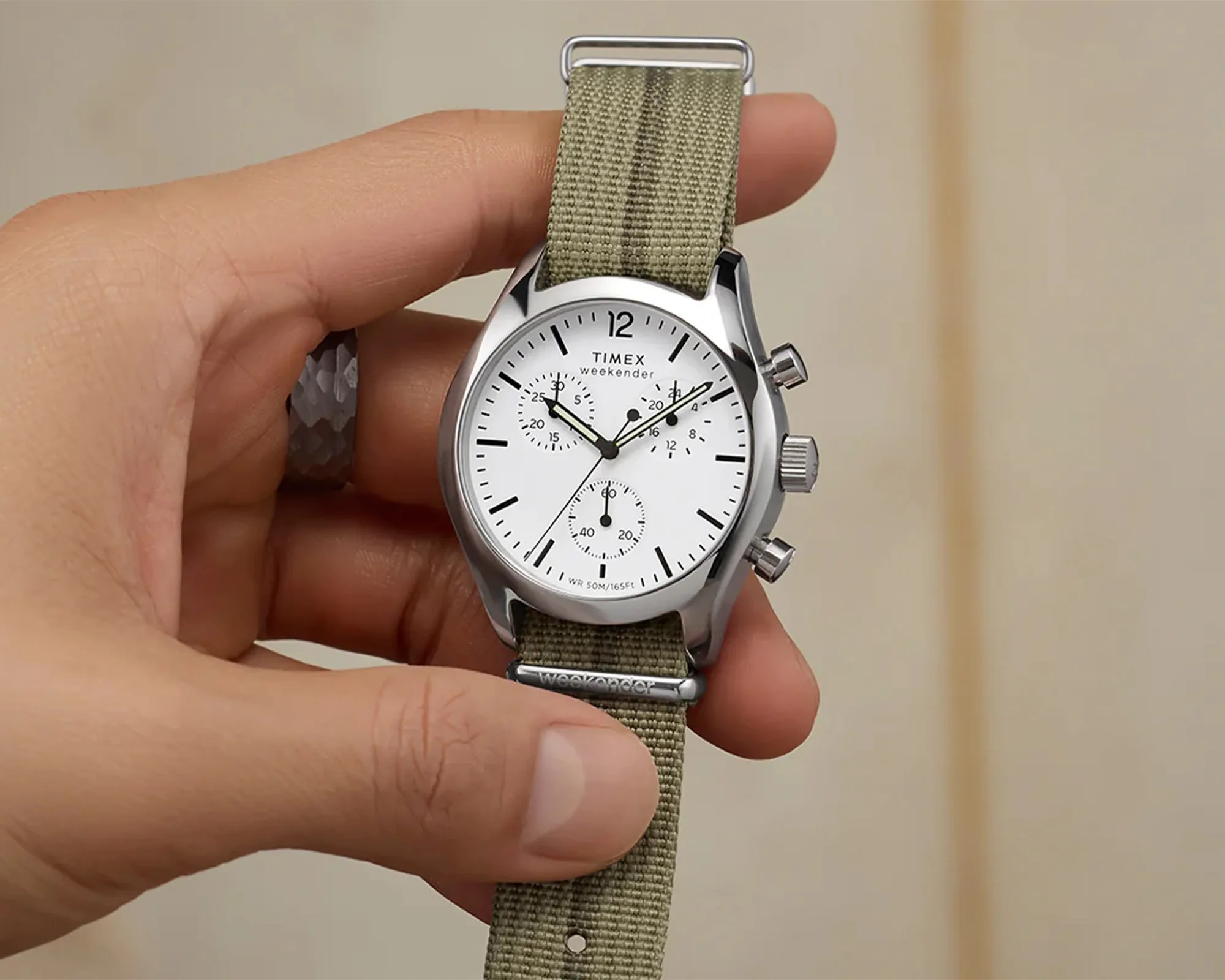 Hand holding a Timex Weekender wristwatch with a white dial, black hour markers, and three subdials. The watch has a silver case and an olive green fabric strap. The watch face shows the time as approximately 10:10:37.