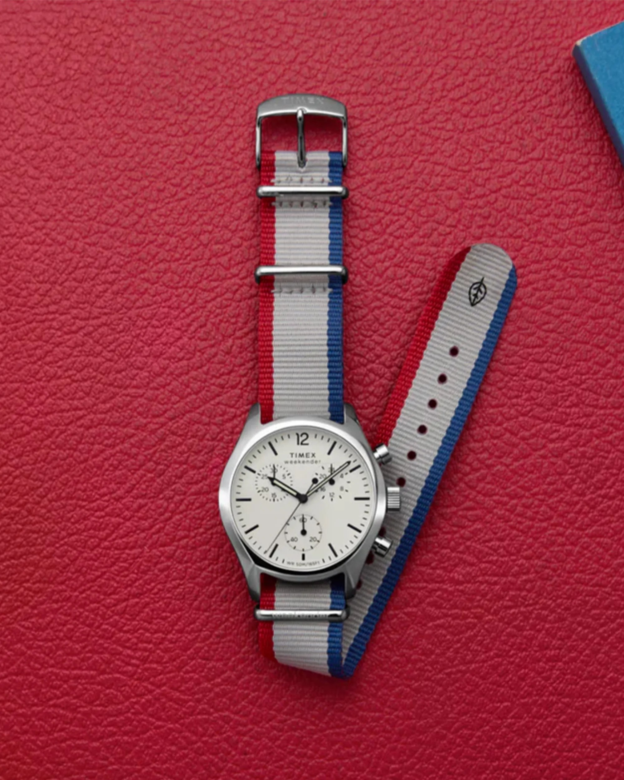 Silver Timex Weekender watch with a white dial and black markings, featuring three subdials and a red, white, and blue striped fabric strap, placed on a textured red surface.