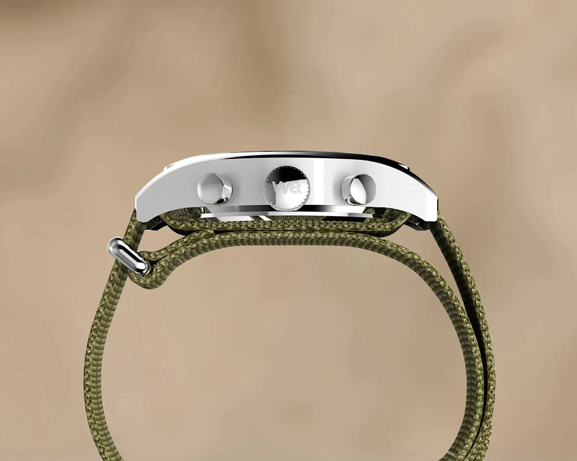 Side view of a wristwatch with a silver metal case and two push buttons flanking a crown engraved with "we." The watch has an olive green woven fabric strap. The background is a smooth beige color.