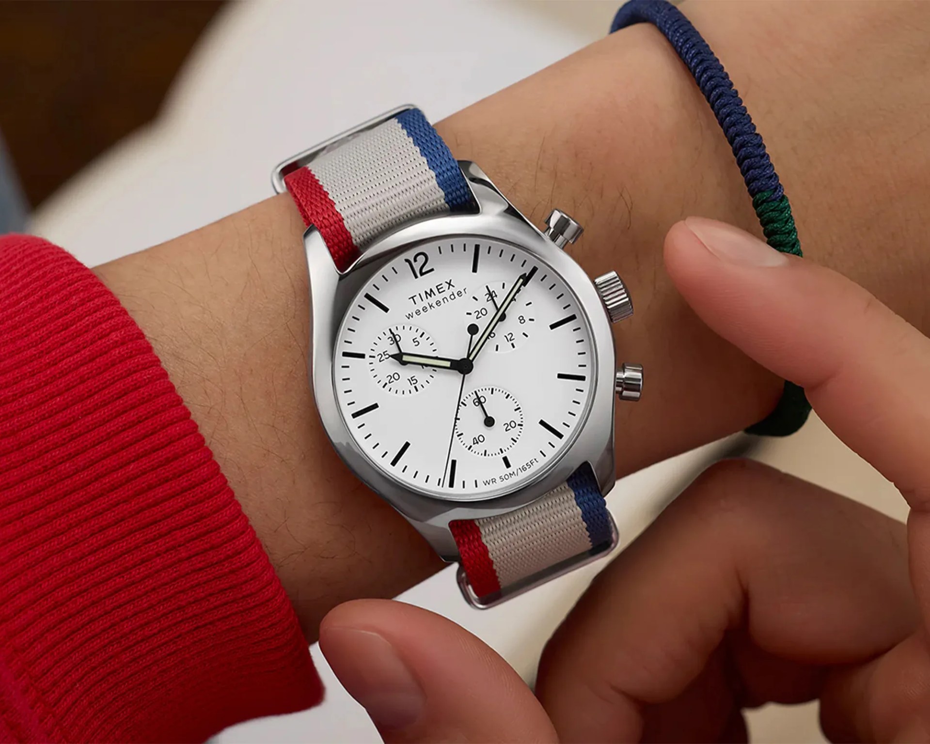 Silver Timex Weekender wristwatch with a white dial, black hour markers, and three subdials. The watch has a fabric strap featuring red, white, and blue stripes. The watch is worn on a wrist with a red sleeve visible, and a hand is adjusting the watch.