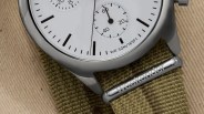 Close-up of a wristwatch with a white dial, black hour markers, and subdials. The watch case is silver, and the strap is olive green fabric with a metal loop engraved with the word "Weekender." The dial also shows "WR 50M/165FT," indicating water resistance.