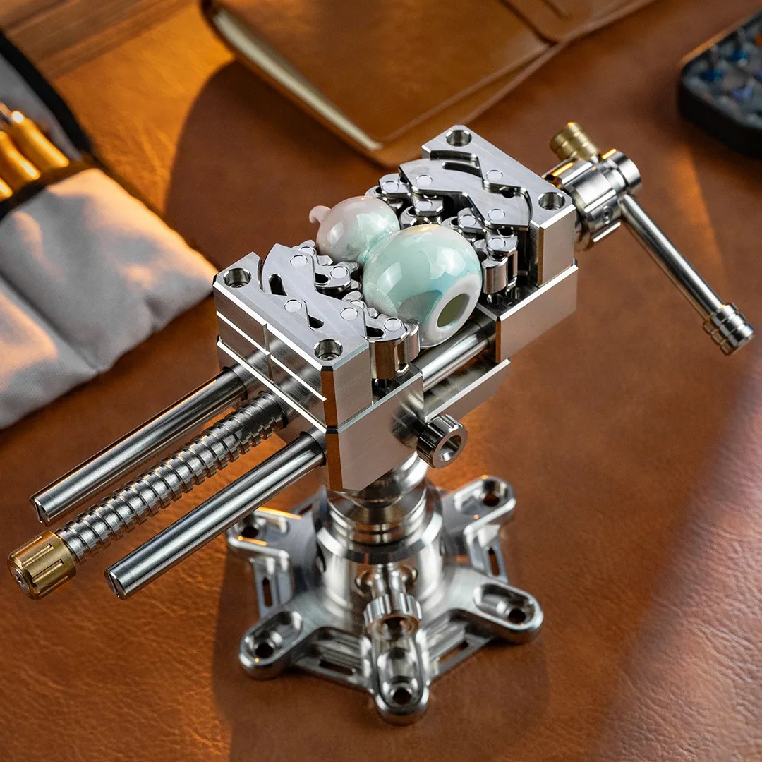 A precision metal vise with a crank handle is holding two polished, spherical stones. The vise is mounted on a sturdy base with multiple screw holes, and the setup is placed on a brown leather surface. In the background, there is a soft pouch with tools and a closed notebook.