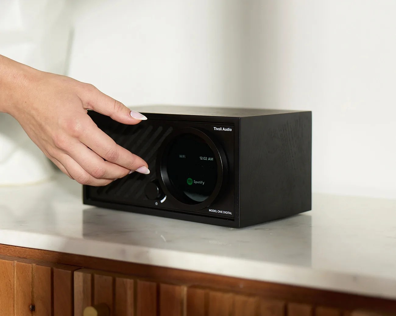 Black Tivoli Audio Model One Digital radio with a circular display showing "WiFi," the time "12:02 AM," and the Spotify logo, placed on a white marble surface with a wooden cabinet below. A hand with manicured nails is adjusting the volume knob on the left side of the radio.