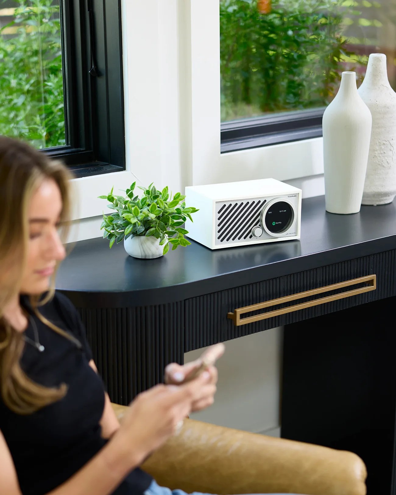 A white Tivoli Audio radio with a circular digital display showing "WiFi" and "Spotify" sits on a black desk with a gold handle. Next to the radio is a small green potted plant in a white marble-patterned pot and two tall white vases. A woman in a black shirt is sitting in a tan chair in the foreground, looking at her phone. The desk is positioned in front of a window with green foliage visible outside.
