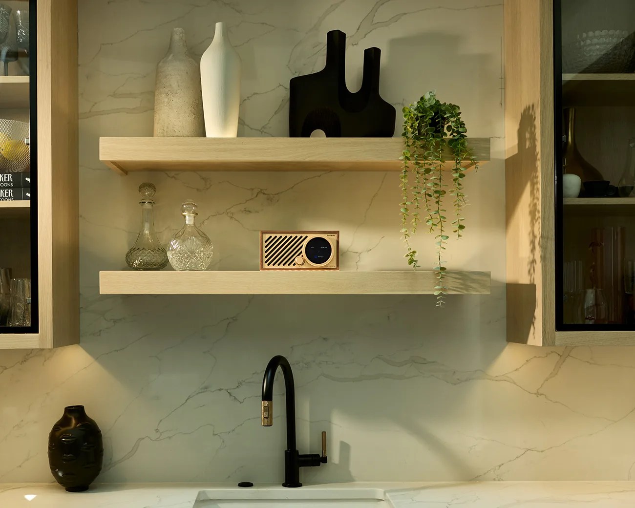 Two light wooden floating shelves mounted on a marble wall above a kitchen sink with a black faucet. The top shelf holds two tall vases, one beige and one white, a black abstract sculpture, and a trailing green plant. The bottom shelf displays two clear glass decanters and a small wooden radio. A black vase sits on the marble countertop to the left of the sink.
