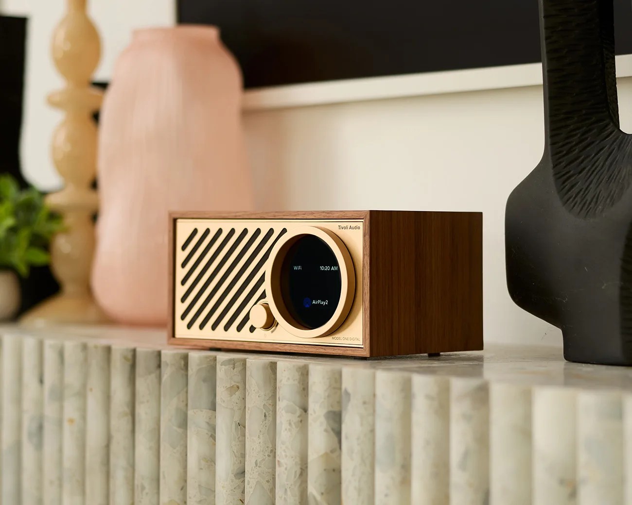 A Tivoli Audio Model One Digital radio with a wooden casing and a gold front panel, featuring a circular digital display showing "WiFi," "10:20 AM," and "AirPlay2," placed on a textured stone surface. The background includes blurred decorative objects in neutral tones.