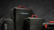 Three black Pelican cases with red handles are shown against a dark textured background. The largest case has the word "PELICAN" printed in light blue on its front. A black Pelican bag with part of the brand name visible is also present on the left side. The cases have a rugged, durable appearance.