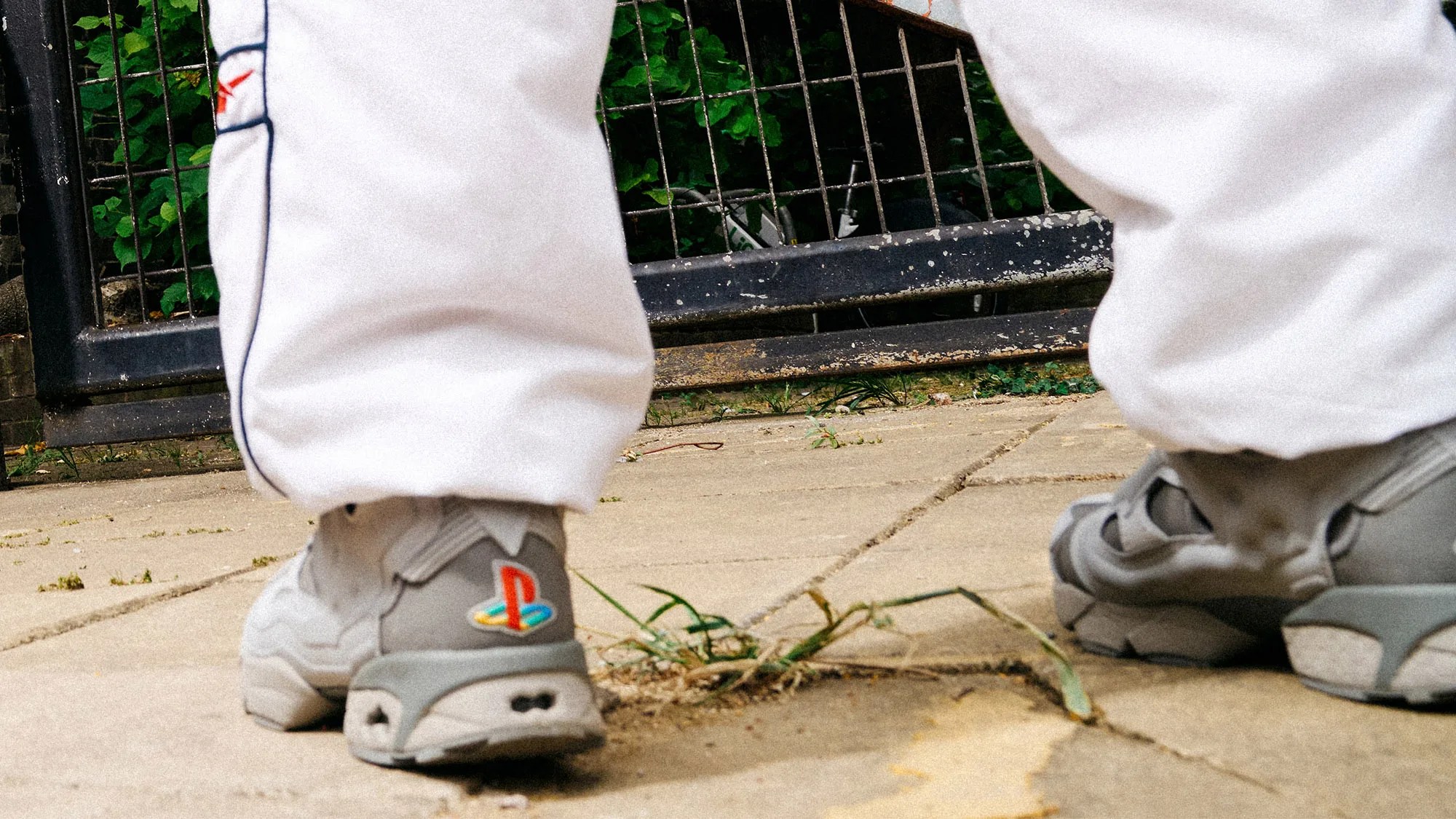 Gray sneakers with a colorful PlayStation logo on the heel, worn with white pants, standing on a paved surface with some grass growing between the cracks. A black metal fence and green foliage are visible in the background.