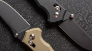 Two folding knives with black blades and textured handles, one with a tan handle and the other with a black handle, placed on a textured black surface.