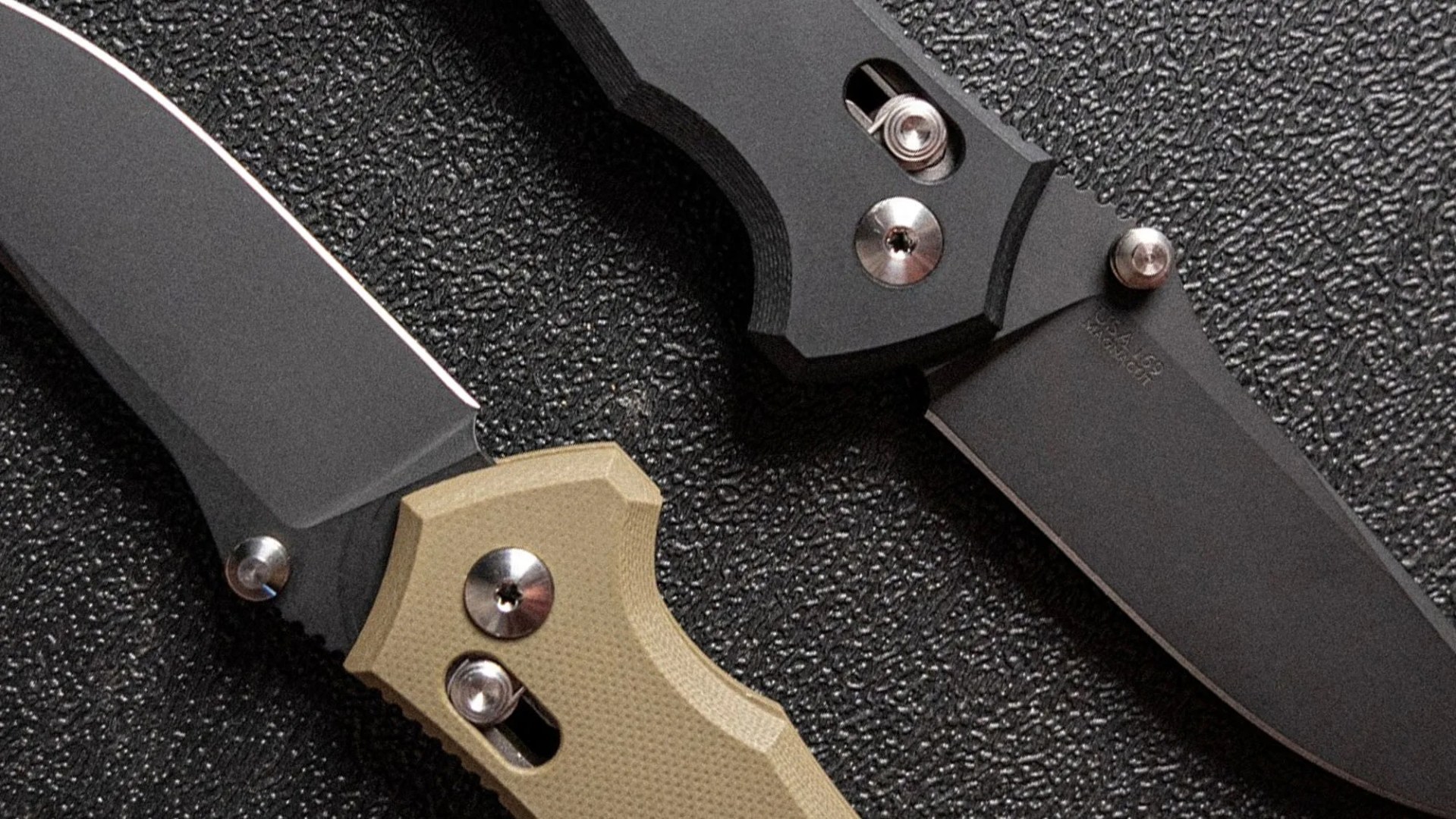 Two folding knives with black blades and textured handles, one with a tan handle and the other with a black handle, placed on a textured black surface.