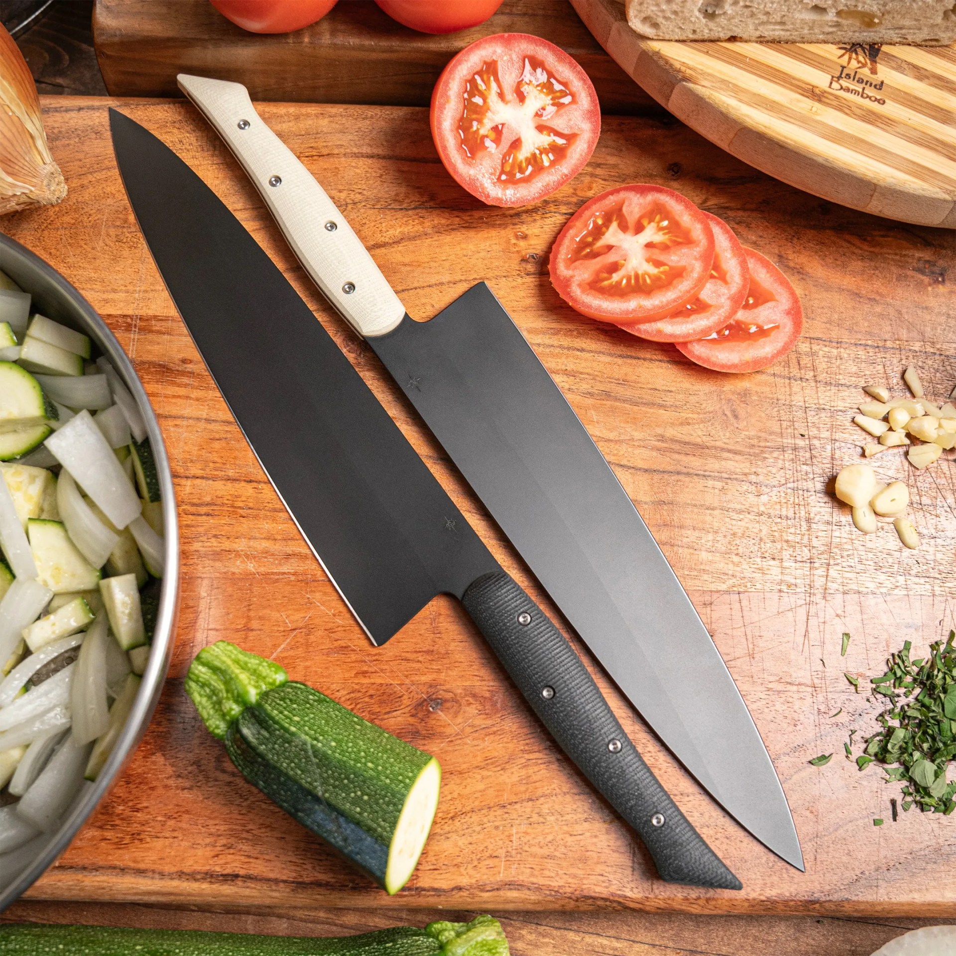 Two black-bladed kitchen knives with textured handles, one white and one black, lie on a wooden cutting board. The board also holds sliced tomatoes, chopped garlic, chopped herbs, a halved zucchini, and a bowl containing chopped onions and zucchini. A round wooden breadboard with a loaf of bread is partially visible in the upper right corner.