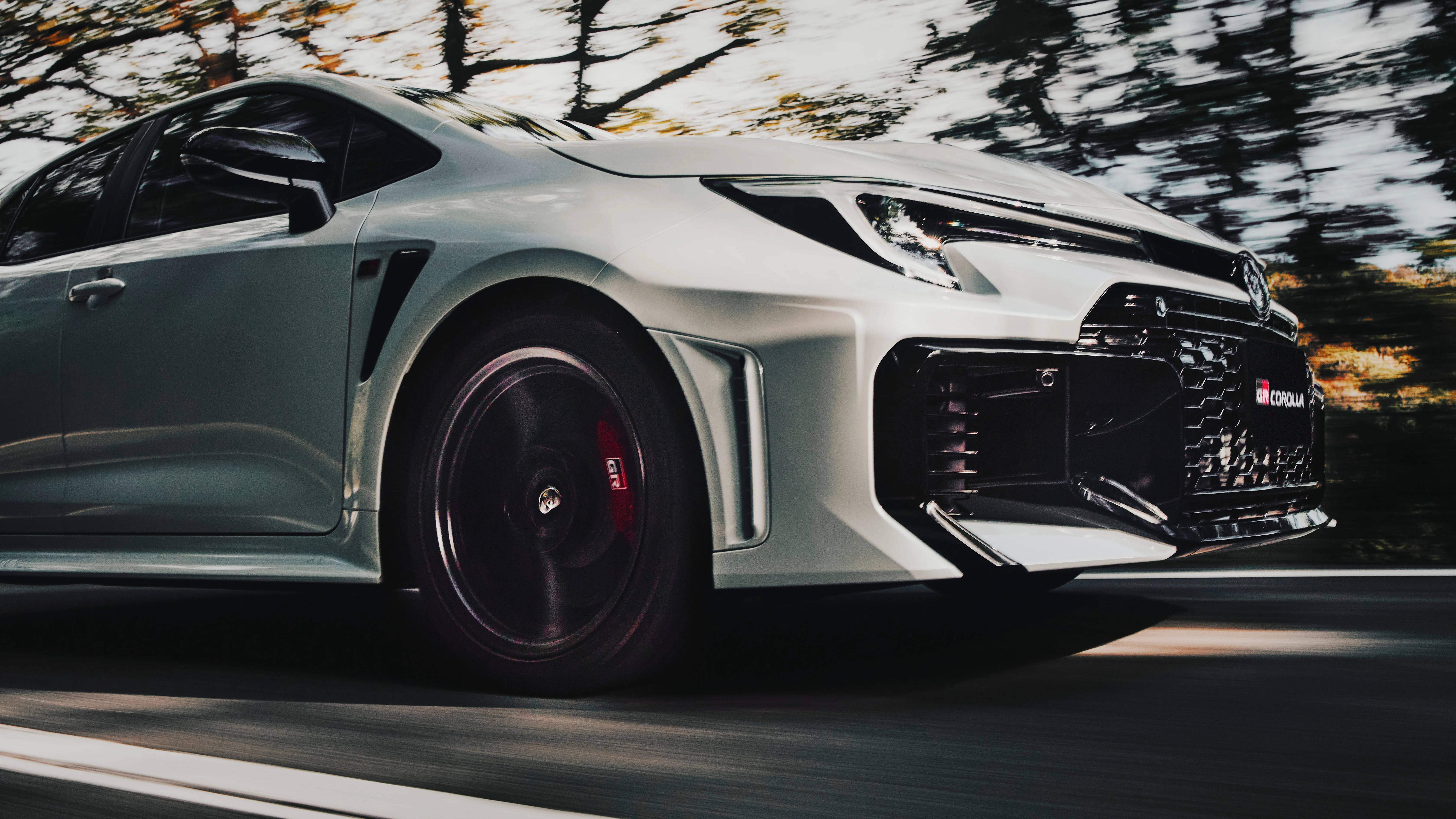 Front side view of a white Toyota GR Corolla in motion, showing the sleek design, black alloy wheel with red brake caliper, and aggressive front grille with GR Corolla badge.