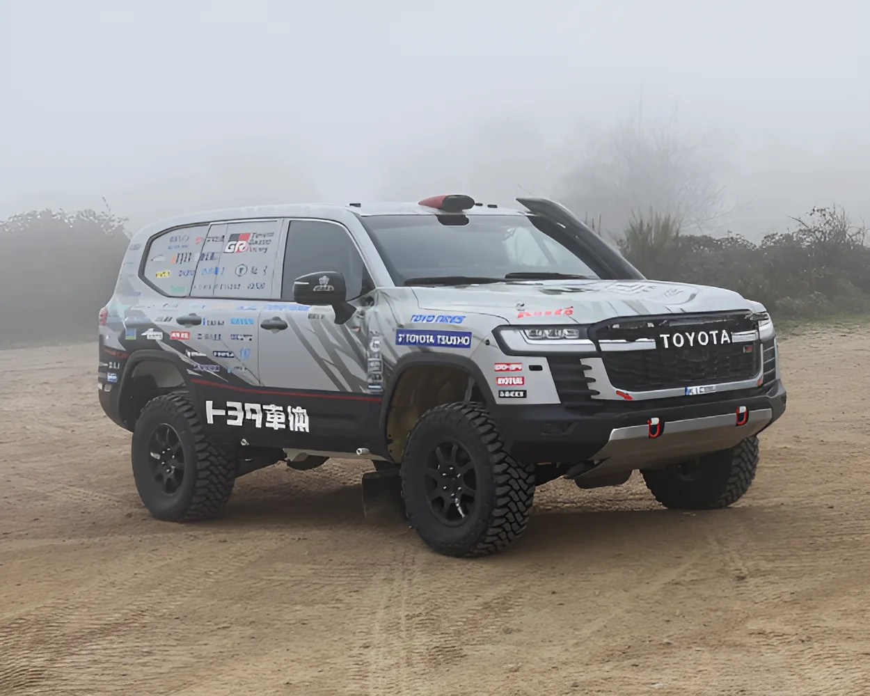 Toyota off-road SUV with a silver and black body, featuring large rugged tires, multiple sponsor decals, and a prominent "TOYOTA" logo on the front grille. The vehicle is parked on a dirt surface with a foggy background.
