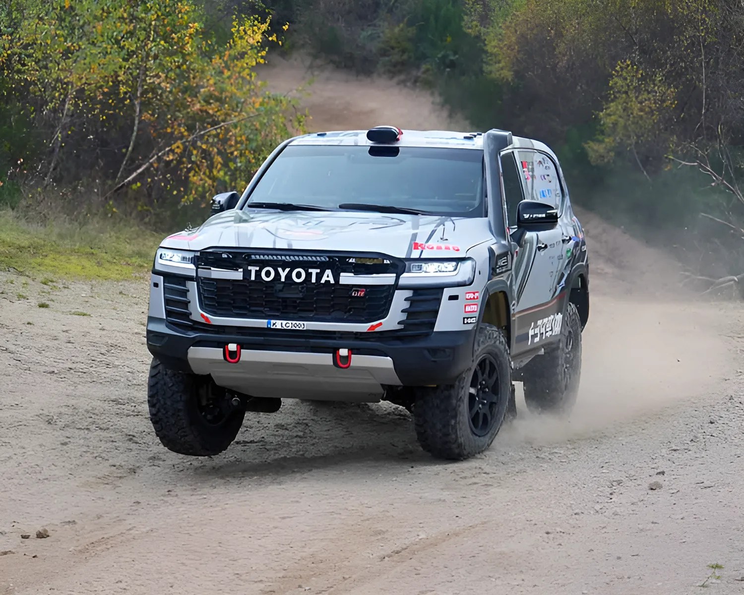 Silver Toyota off-road SUV driving on a dirt trail, kicking up dust, with large rugged tires and a black grille featuring the Toyota logo. The vehicle has red tow hooks on the front bumper and various decals on the sides and hood. Trees and greenery are visible in the background.