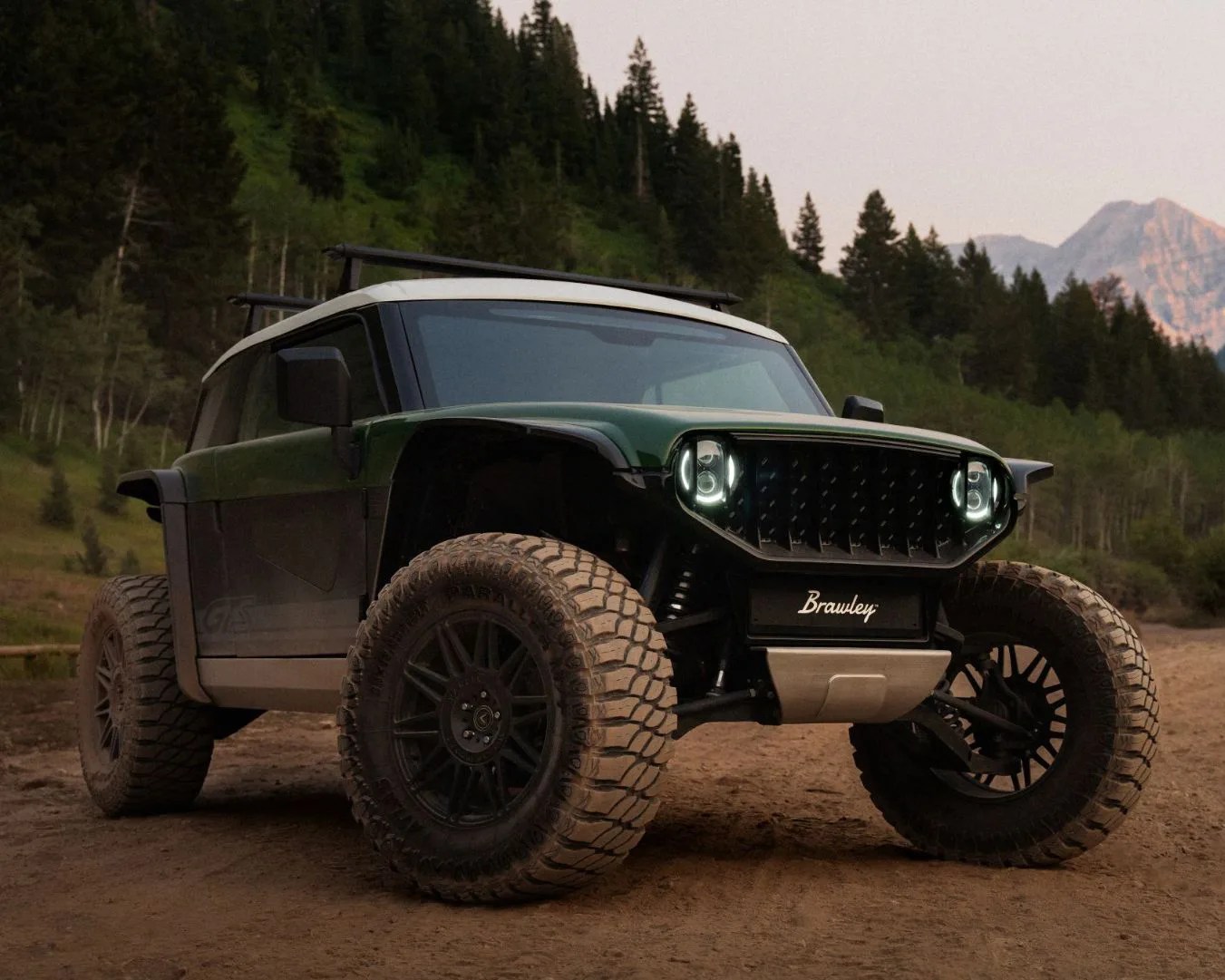 A rugged off-road vehicle with large, knobby tires and a dark green body with black accents. It has a white roof with roof racks and distinctive round LED headlights. The front grille is black with a textured pattern, and the license plate area displays the name "Brawley." The vehicle is parked on a dirt path with a forest and mountains in the background.