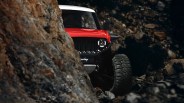 A red and white off-road vehicle with large tires is partially visible behind a rocky outcrop, navigating a rugged, rocky terrain. The vehicle's front grille and one headlight are clearly seen, with the headlight illuminated. The background is dark and filled with rocks.