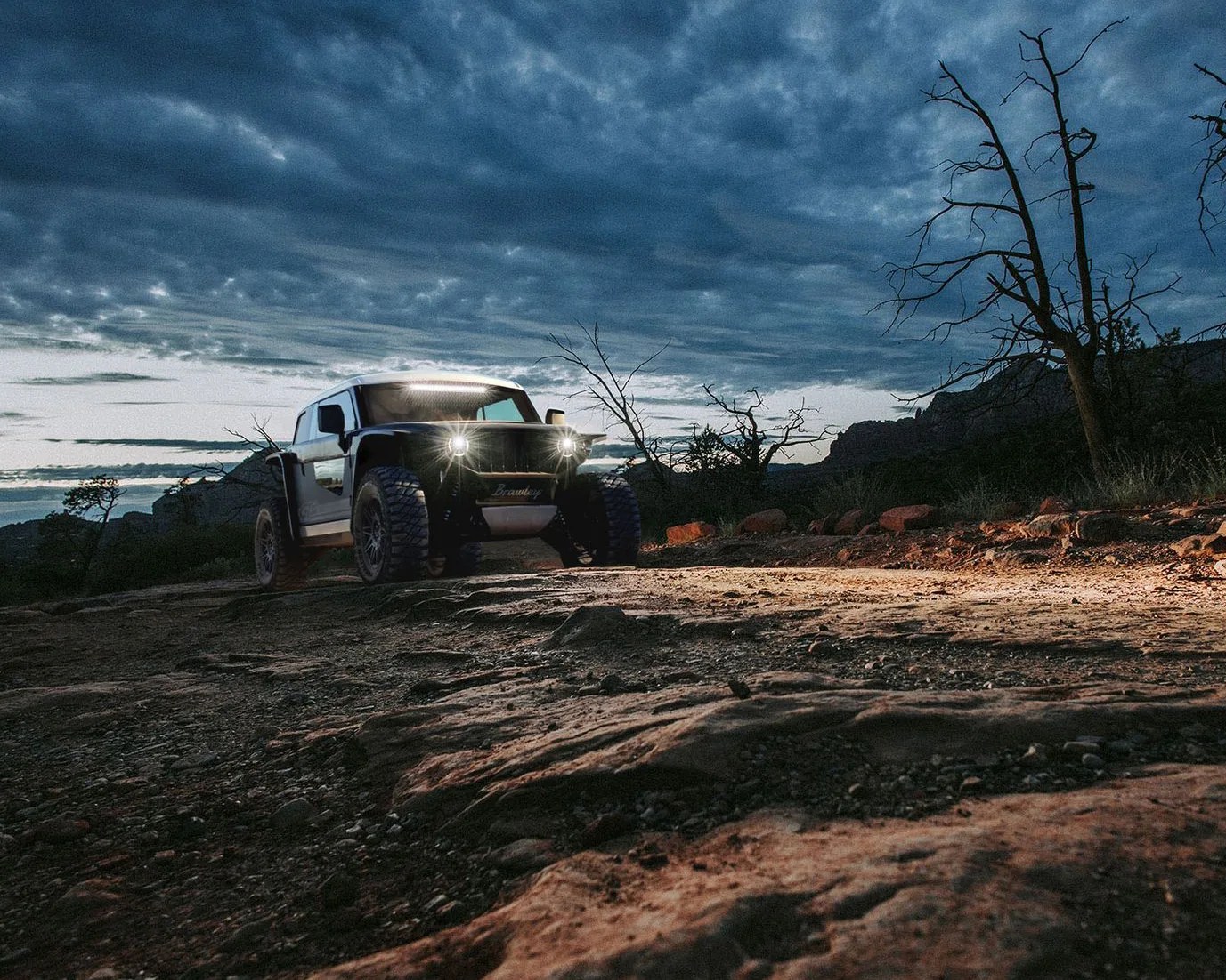 A rugged off-road vehicle with bright headlights and a light bar on the roof is parked on a rocky dirt trail at dusk. The sky is filled with dark, dramatic clouds, and there are leafless trees and rocky terrain surrounding the vehicle.