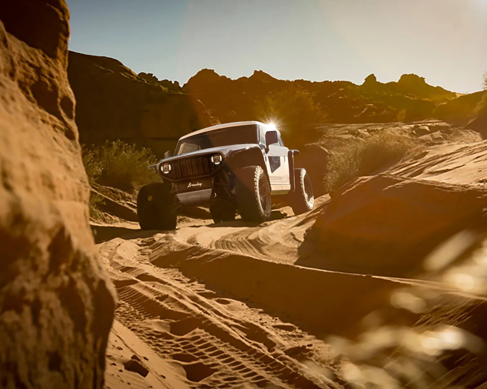 A rugged off-road vehicle with large tires driving on a sandy, rocky trail in a desert landscape during sunset, with sunlight glinting off the vehicle's roof. The vehicle has a distinctive front grille and the word "Brawley" visible on the front.