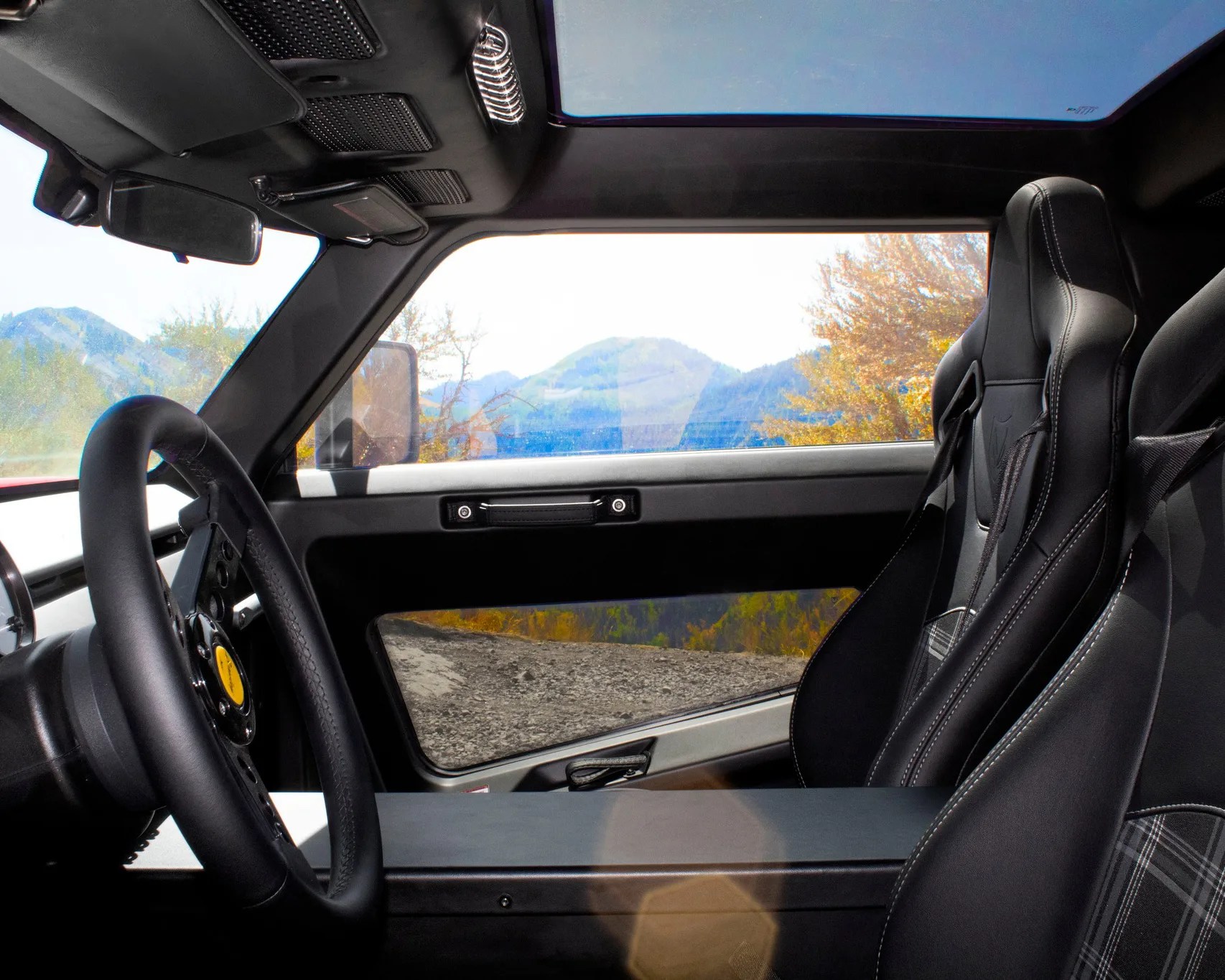 Interior of a vehicle featuring black leather bucket seats with white stitching and plaid fabric inserts, a black steering wheel with a yellow center emblem, and a large panoramic sunroof. The door panel is black with a simple handle, and the window shows a scenic view of mountains and autumn trees outside.
