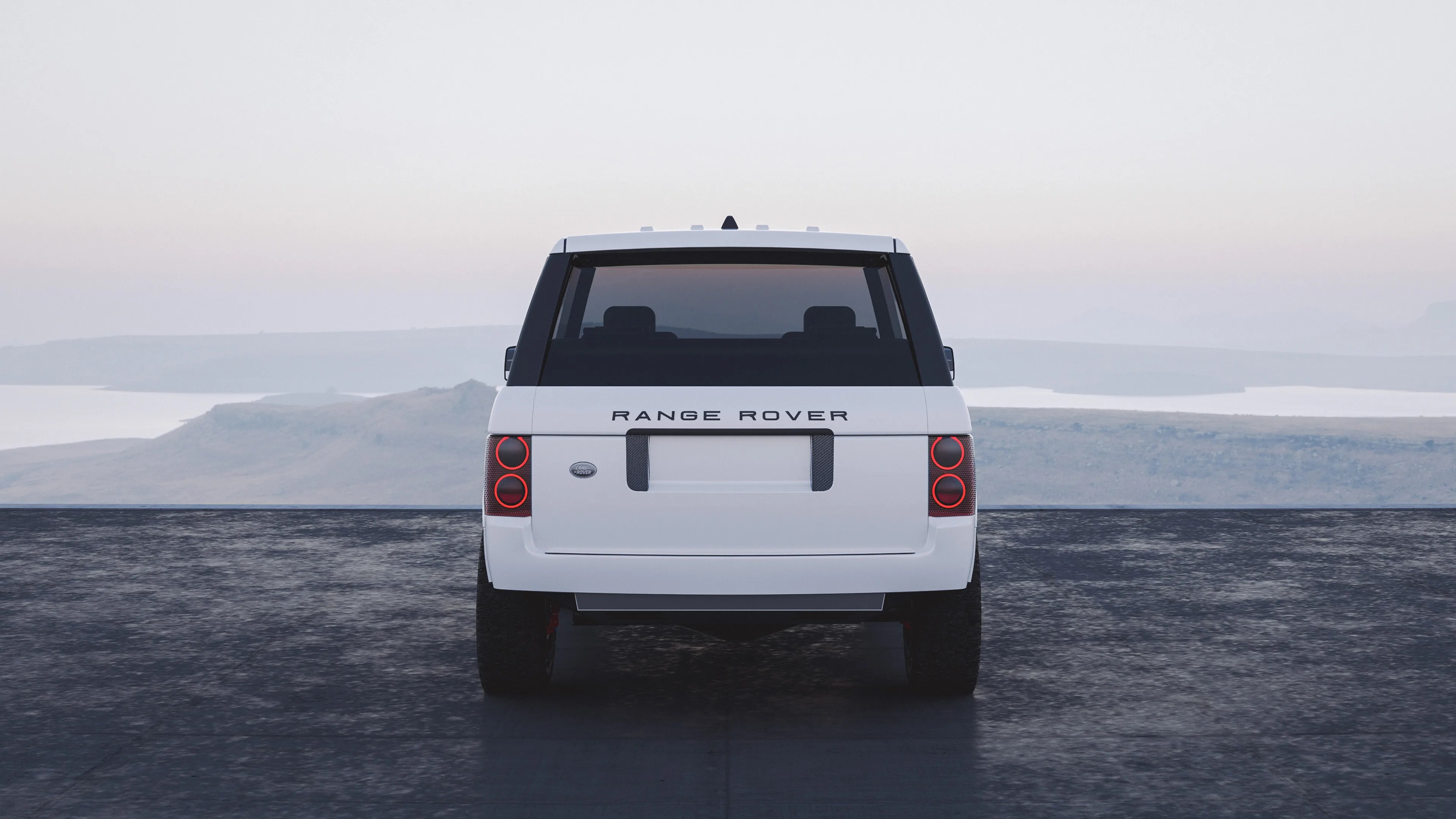 White Range Rover viewed from the rear, with distinct circular red tail lights and tinted rear window, parked on a dark surface with a hazy mountainous landscape in the background.