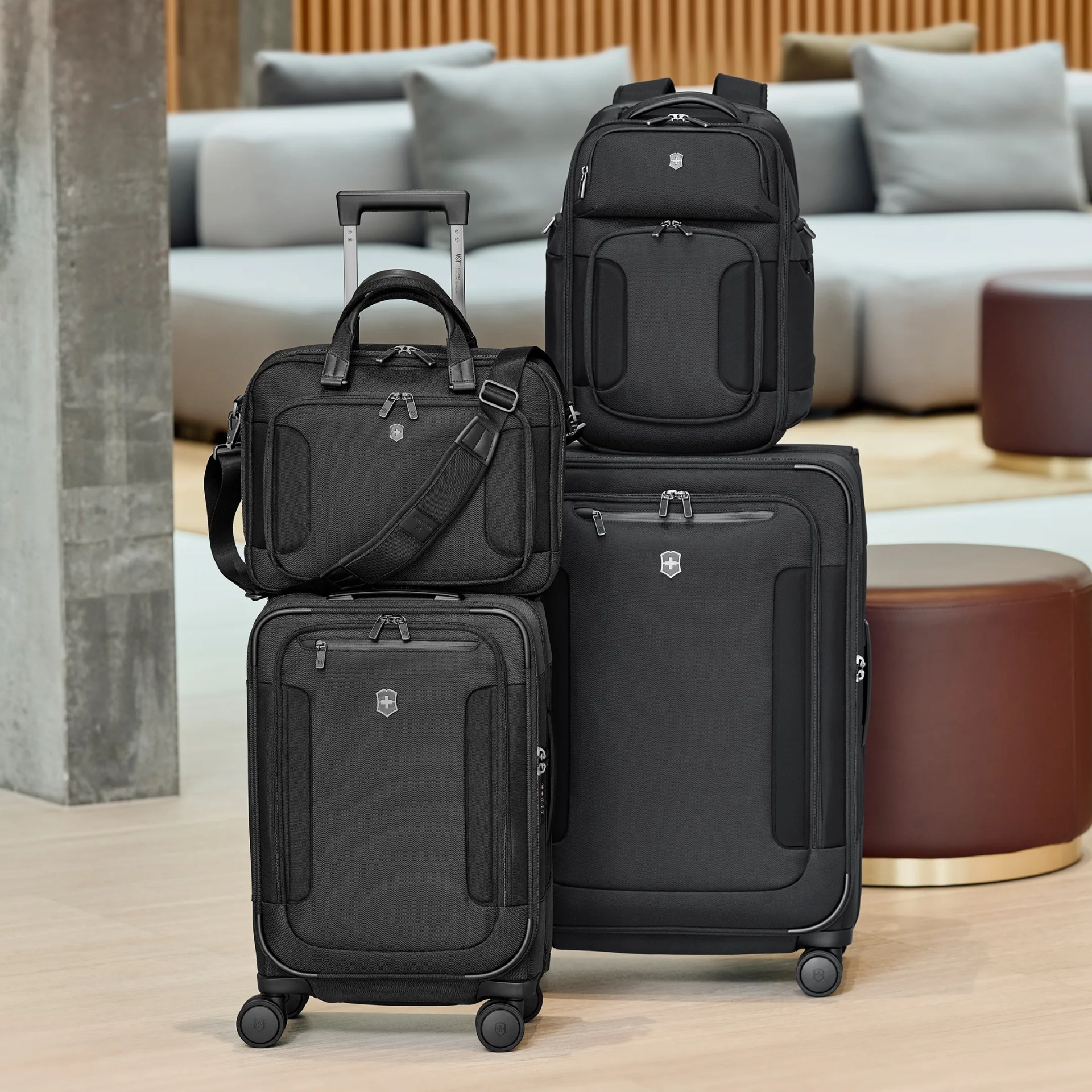 A set of four black luggage pieces including two upright suitcases with wheels, a backpack, and a wheeled briefcase with a shoulder strap, all featuring a small silver emblem on the front. The luggage is displayed indoors on a light wood floor with a modern sofa and round ottoman in the background.