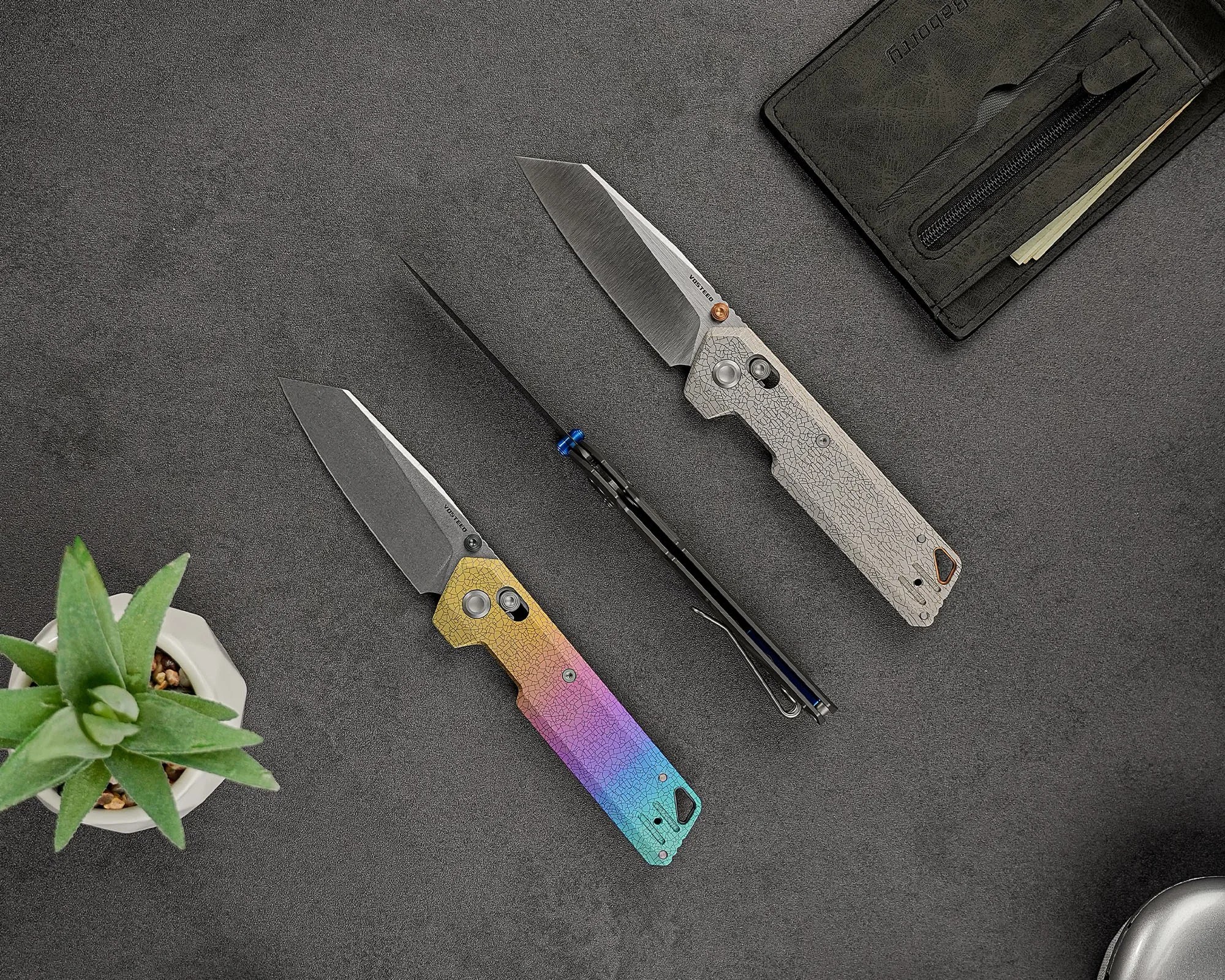Two folding knives with textured handles and straight-edged blades are displayed on a dark surface. One knife has a gradient handle transitioning from yellow to purple to blue, while the other has a light gray handle with a cracked pattern. A small green succulent plant in a white pot is positioned in the bottom left corner, and a black wallet with a zipper pocket is partially visible in the top right corner.
