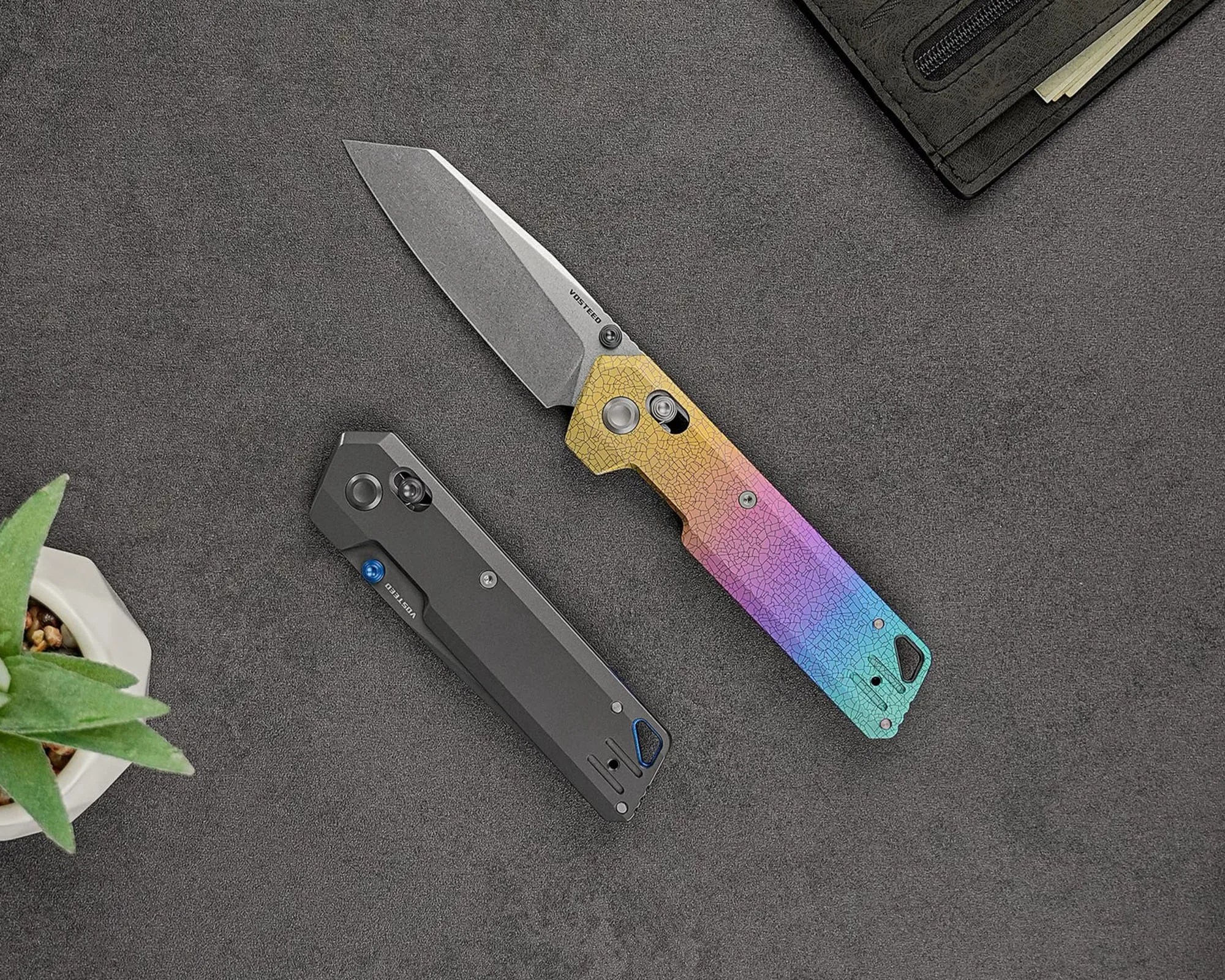 Two folding knives are displayed on a dark gray textured surface. One knife is open, featuring a blade with a stonewashed finish and a handle with a gradient color pattern transitioning from yellow to purple to blue, with a cracked texture design. The other knife is closed, with a matte black handle and a blue thumb stud. A small green succulent plant in a white pot is partially visible in the bottom left corner, and a black wallet with some cash is partially visible in the top right corner.