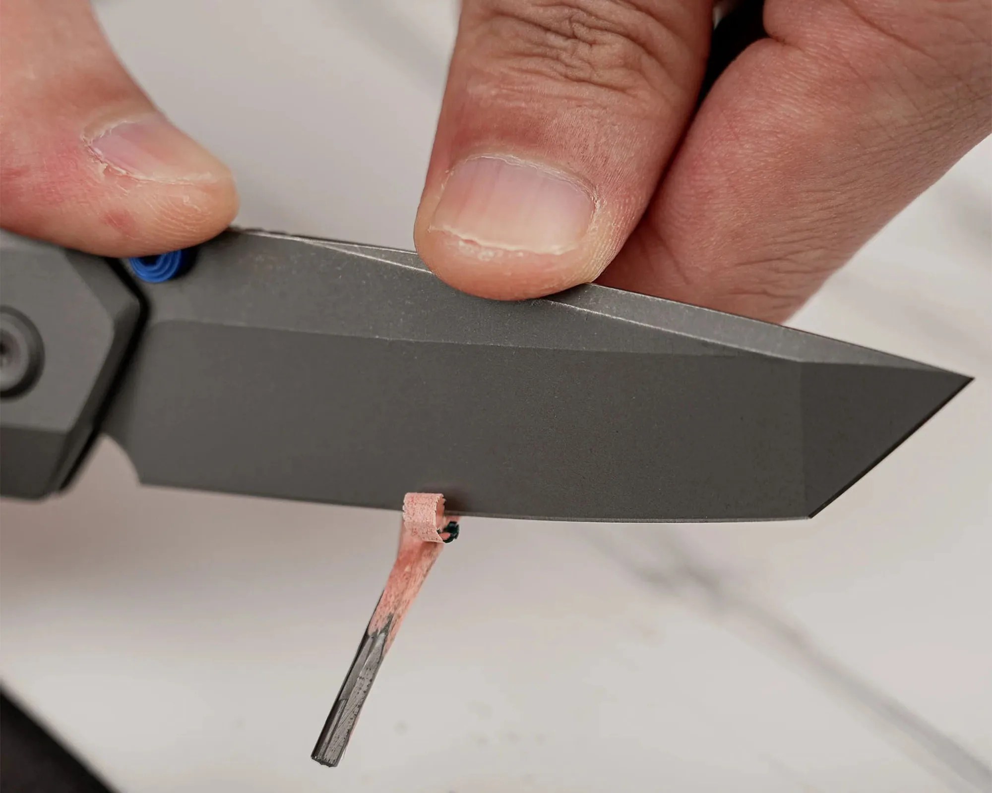 A close-up of a hand holding a black folding knife blade with a small curled shaving of a pink sharpening stone being applied to the blade edge. The sharpening stone is held against the blade to hone the edge.