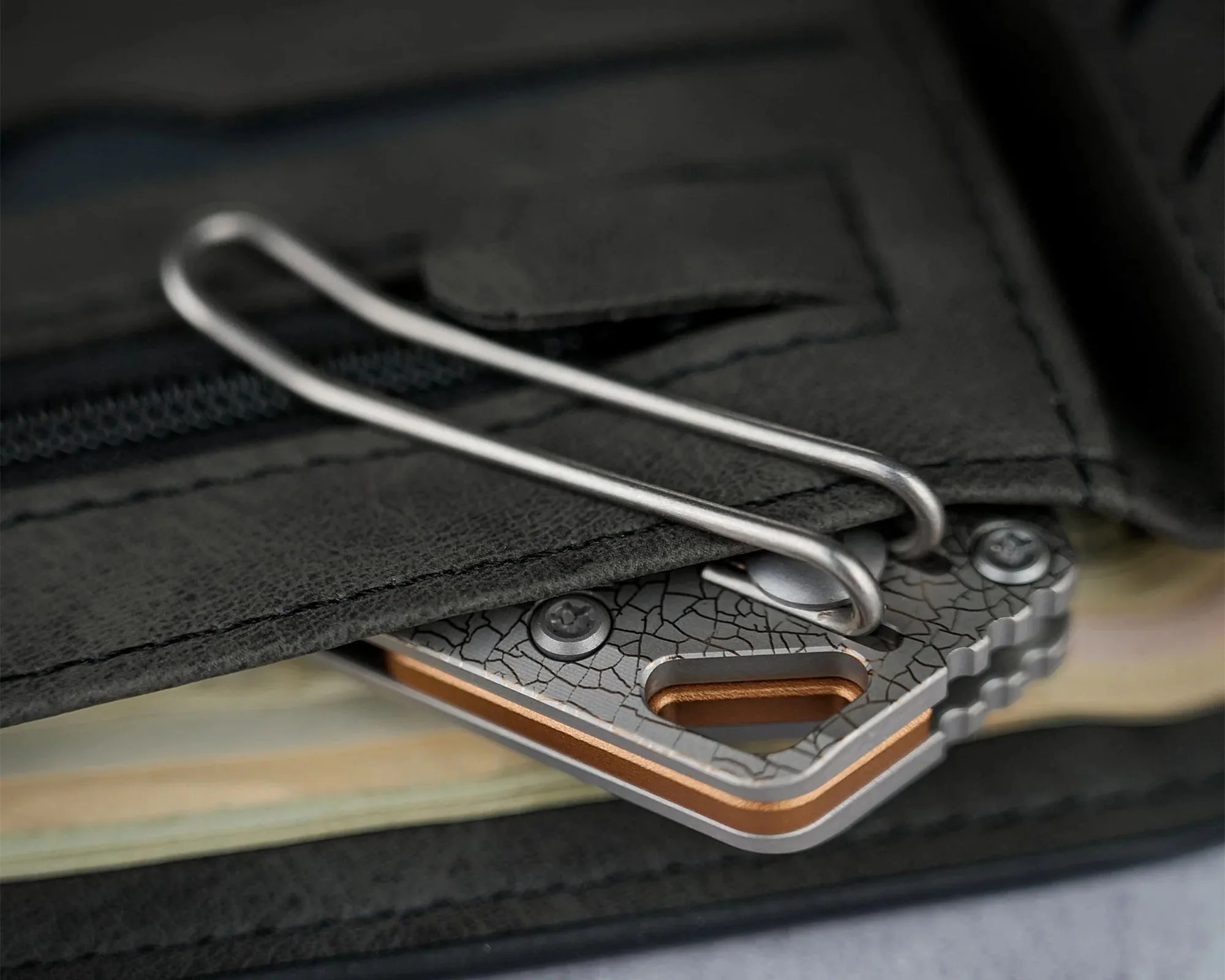 Close-up of a textured metal money clip attached to a black leather wallet, holding folded cash. The clip has a cracked pattern design and is secured with two screws.