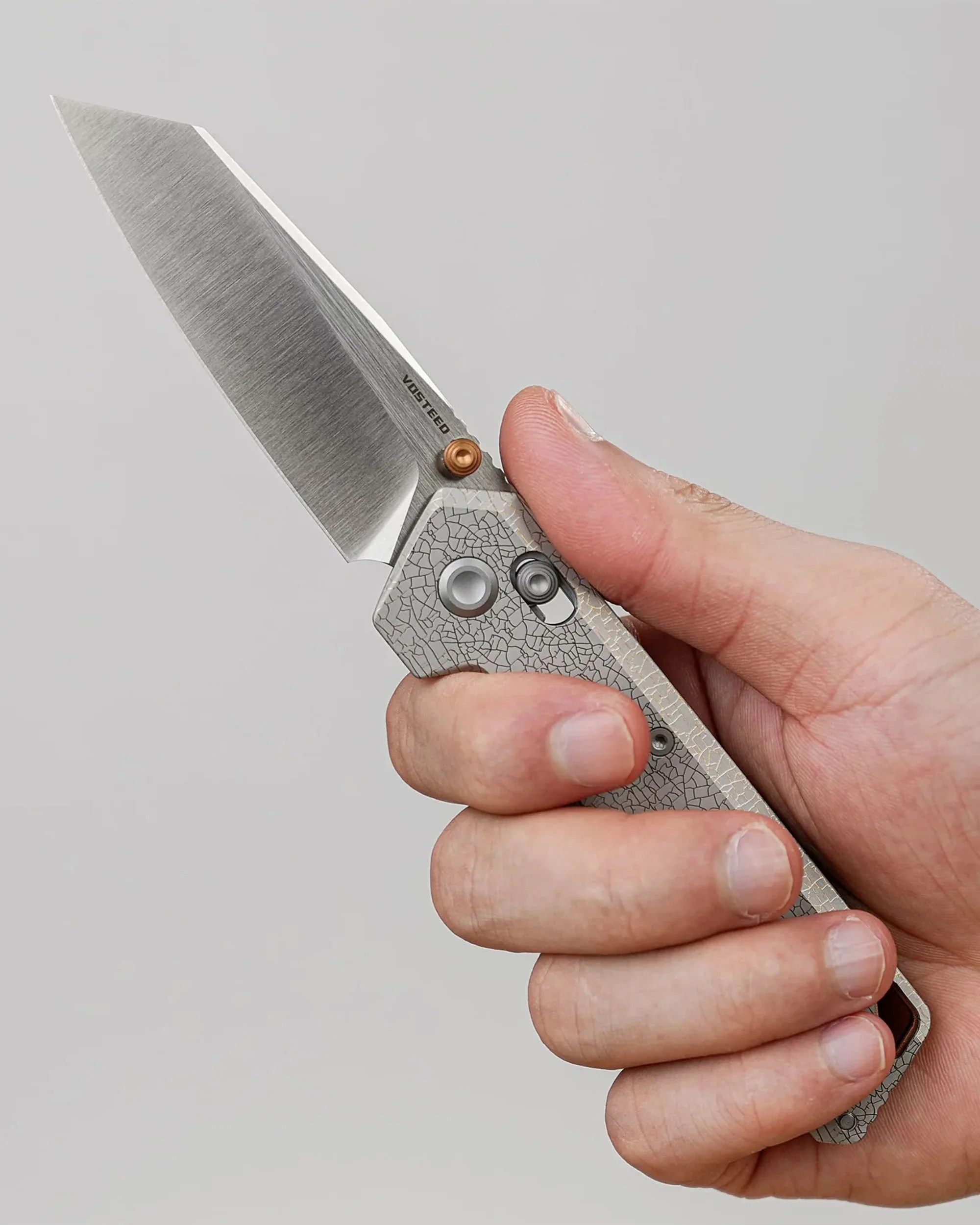 Hand holding a folding knife with a textured gray handle and a wide, straight-edged stainless steel blade. The blade has a brushed finish and the word "VOSTEED" printed near the pivot. The handle features a circular pivot and a small sliding lock mechanism.