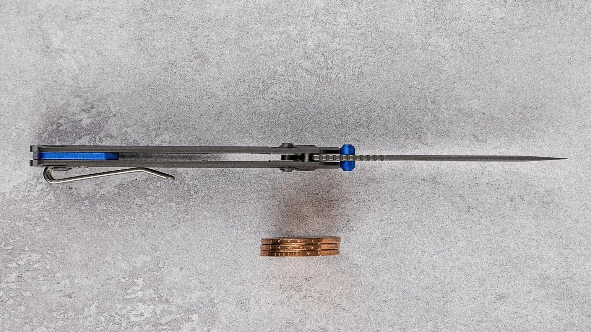 Slim folding knife viewed from the top, showing a thin, pointed blade and a blue handle liner with a metal pocket clip. Three stacked copper coins are placed below the knife for scale. The background is a textured gray surface.