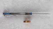 Slim folding knife viewed from the top, showing a thin, pointed blade and a blue handle liner with a metal pocket clip. Three stacked copper coins are placed below the knife for scale. The background is a textured gray surface.