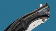 Close-up of a folding knife handle featuring a textured black grip with a subtle swirling pattern, a blue and bronze circular pivot, a small black screw, and a brushed metal section with an engraved signature. The background is solid blue.