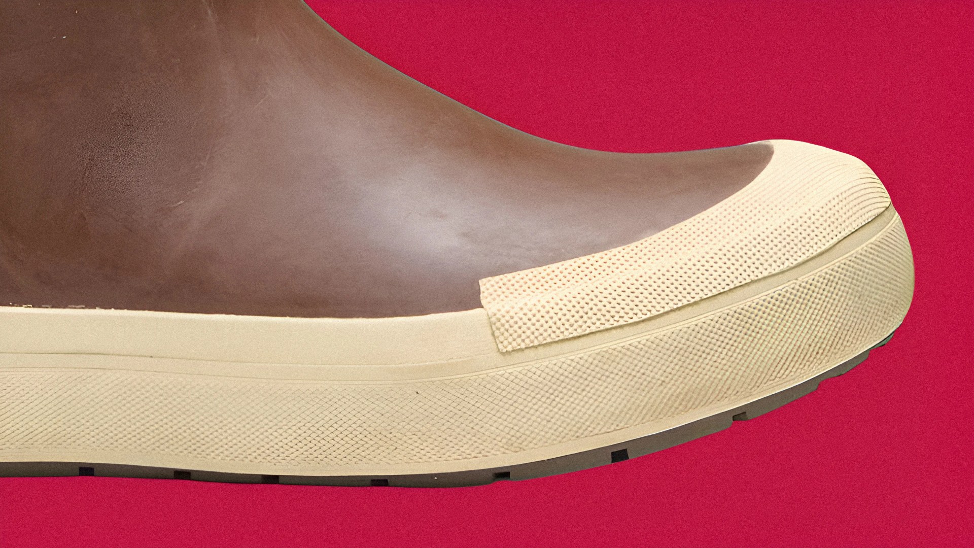 Close-up of the front part of a brown rubber boot with a beige textured toe cap and sole, set against a red background.
