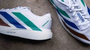 White athletic shoes with blue, green, and brown geometric accents. The left shoe shows the text "Humanrace" near the heel and "LIGHTSTRIKE PRO" on the sole. The right shoe has white laces with a tag labeled "ADIZERO." The shoes are placed on a gray concrete surface.